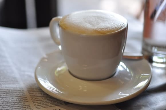 Life in Italy vs. Life in France: Is coffee better in France or Italy? We love Italian Cappuccino, like this frothy one in a white cup and saucer on a newspaper laid out on a table.