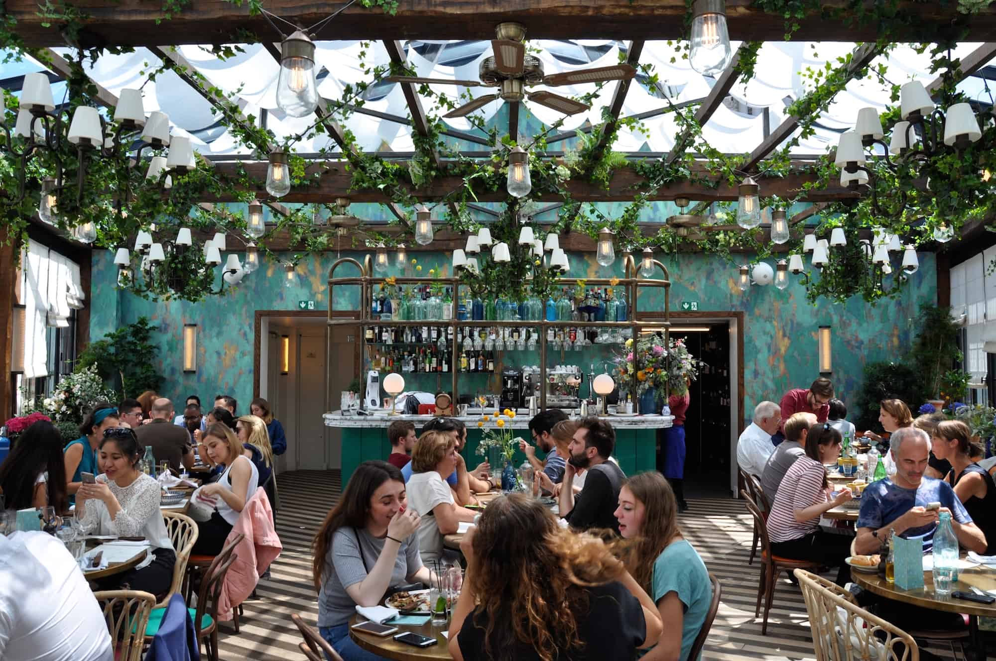 The Big Mamma Group offers Trendy, Fresh, Authentic Italian Dining in Paris at several locations including Pink Mamma in South Pigalle, where there are tables under a glass roof adorned with plants.