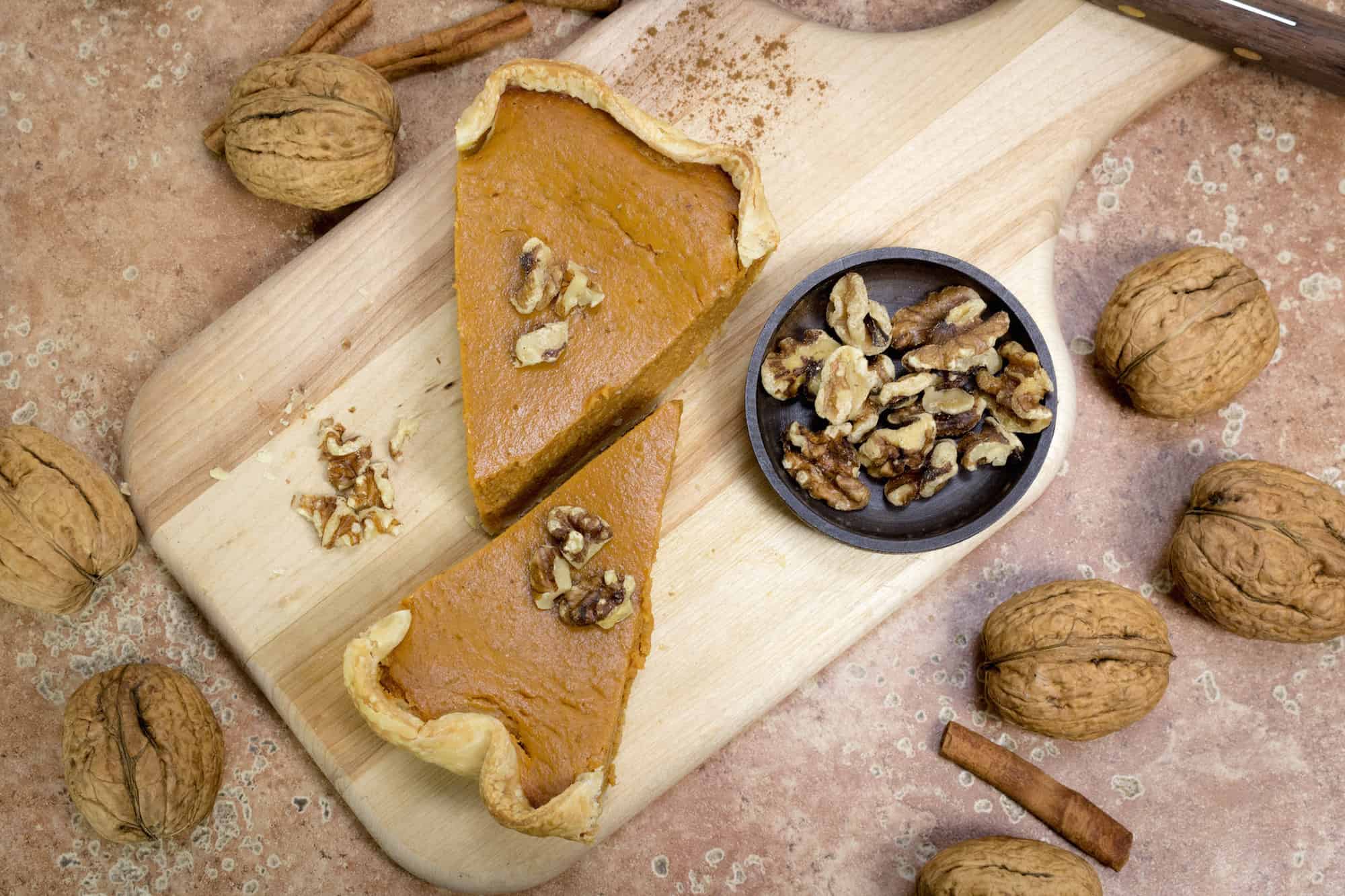 Two slices of yellow pumpking pie sits on a wooden board with a small bowl of walnuts.