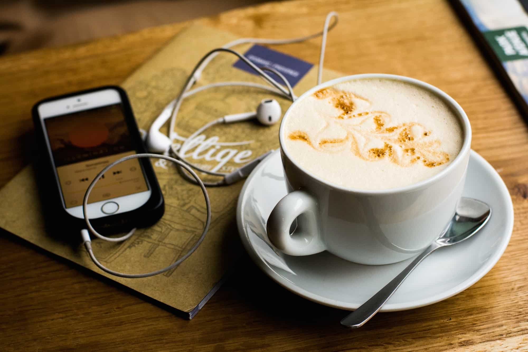 HiP Paris Blog rounds up the top French podcasts to enjoy while you have a coffee, like this creamy Cappuccino on a wooden table, next to a phone with a podcast on the screen.