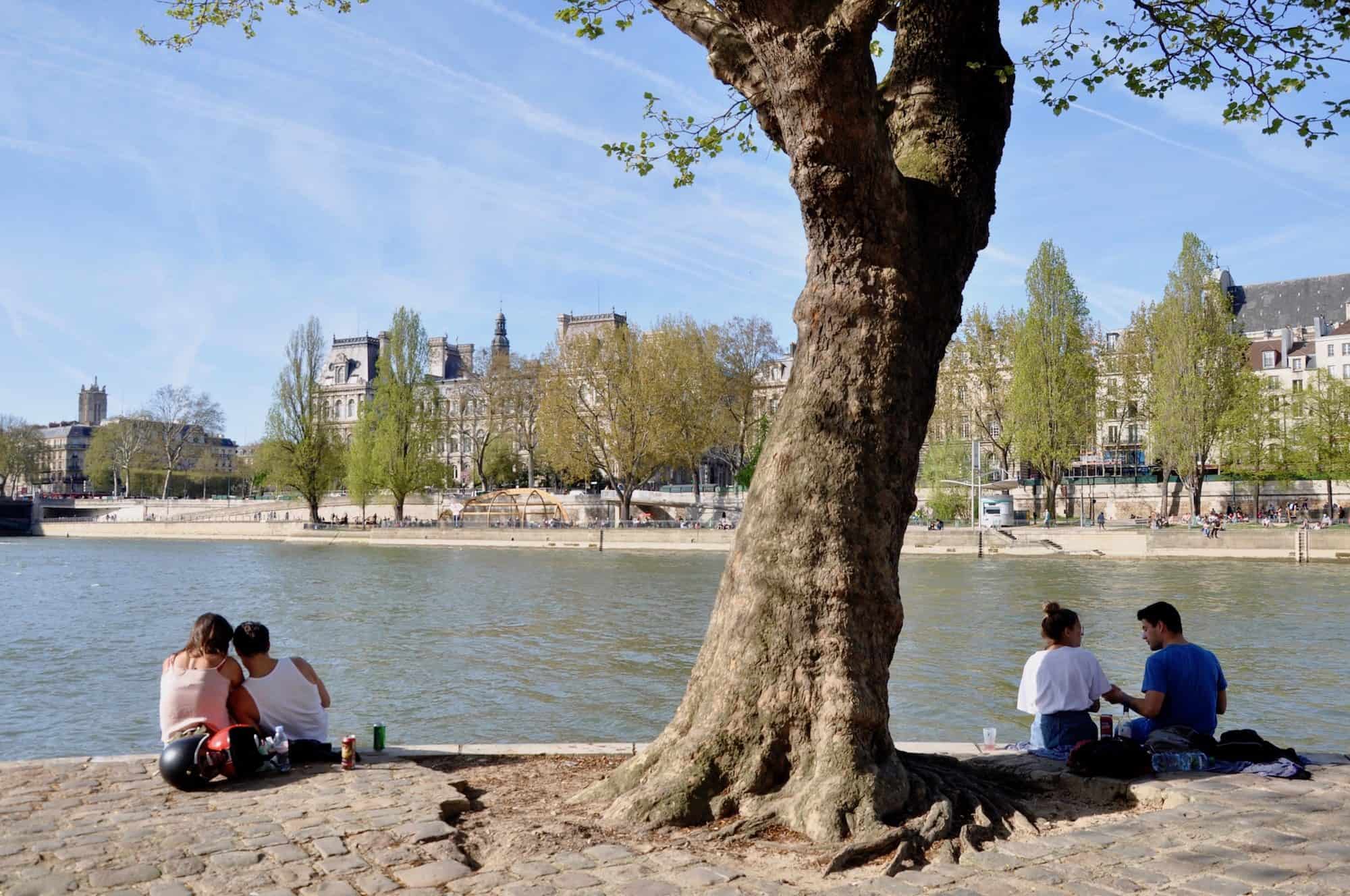 Learning French in Paris is best in summer when you can meet people to practise at picnics on the River Seine out in the sunshine like these two couples sitting on the banks facing the Haussmann houses on the other side.