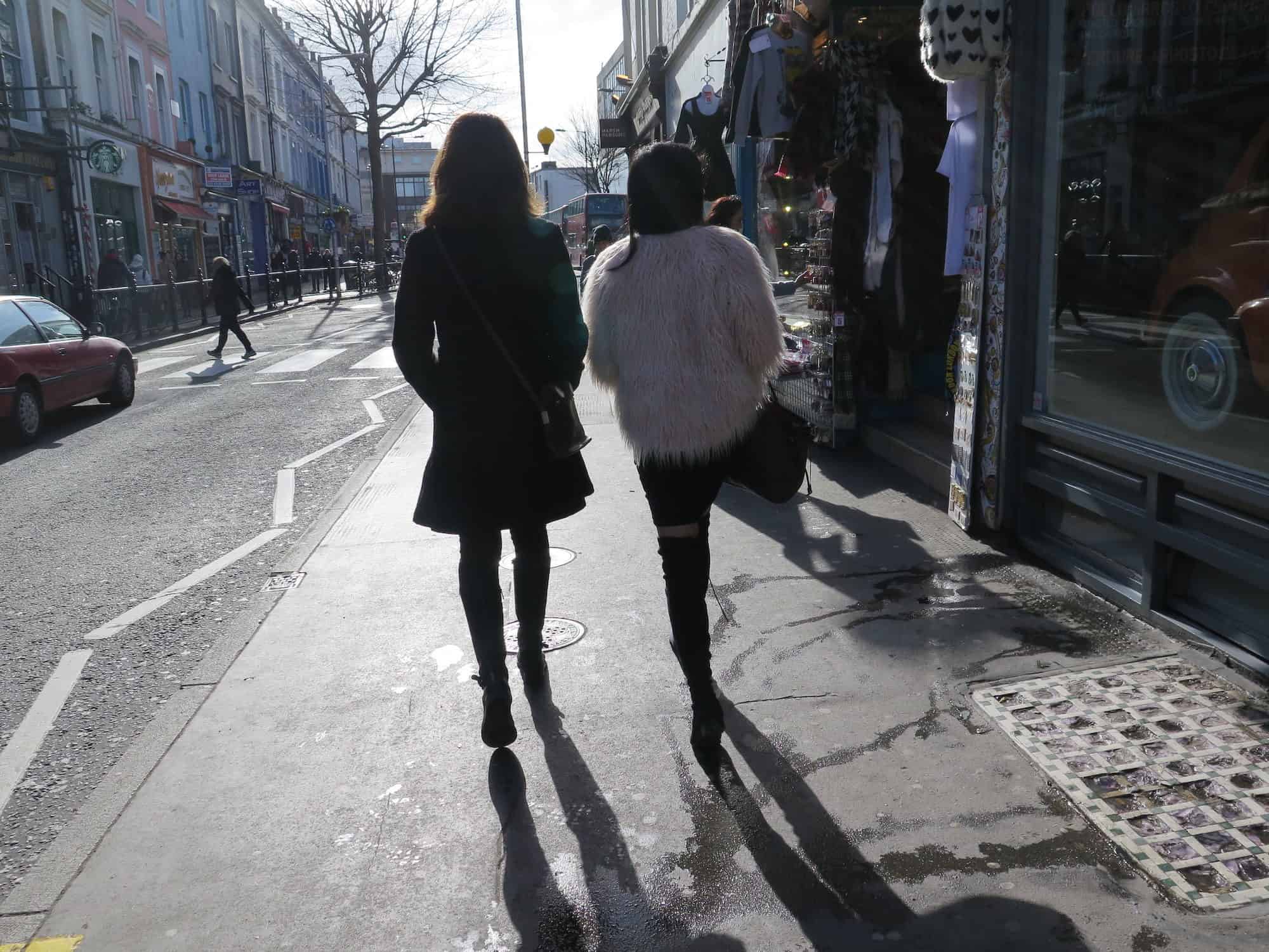 Two girls walking along a street in the London sunshine, one of them wears a fluffy pink jacket.