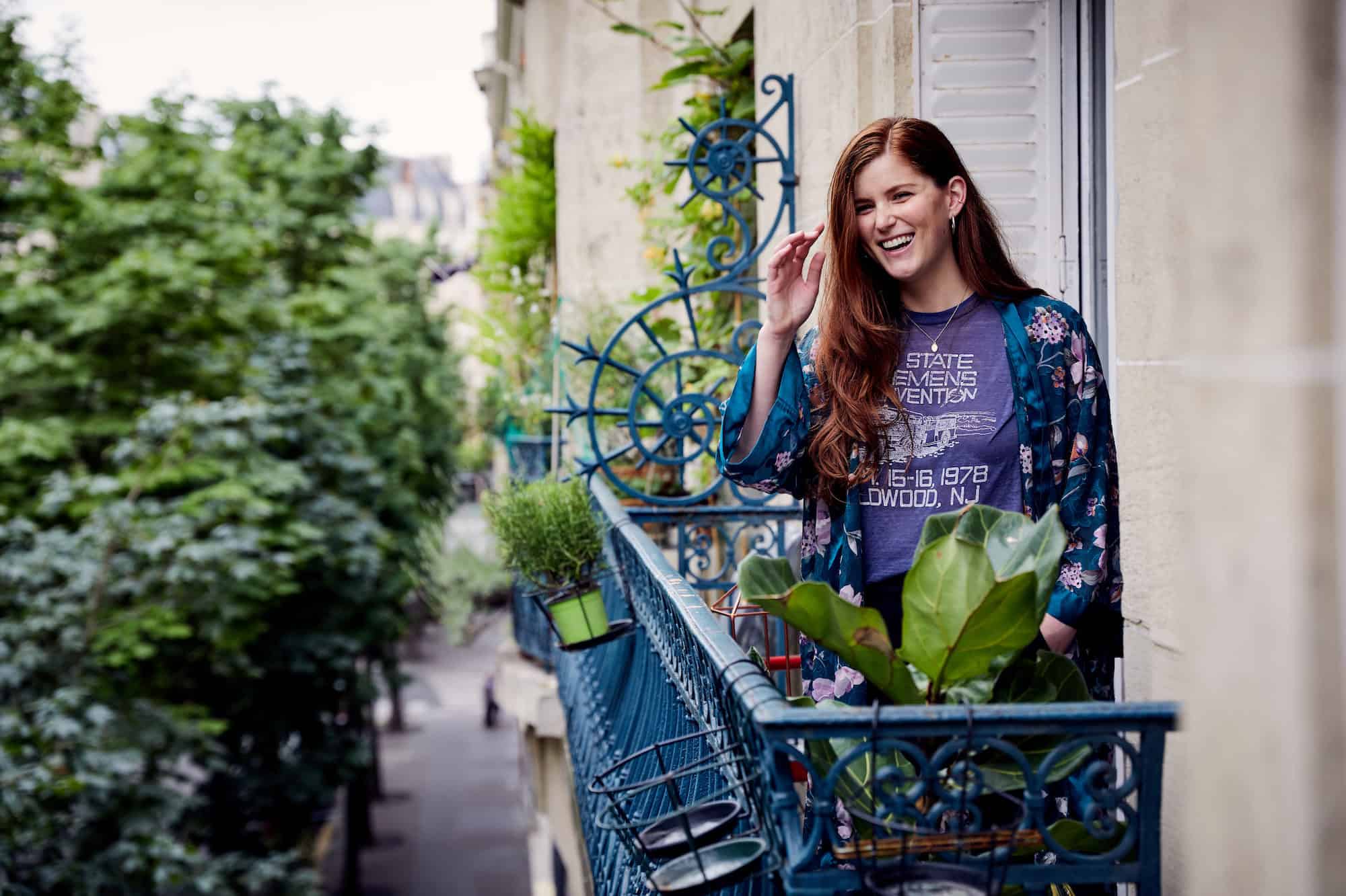 A woman living in Paris and laughing, while standing on her balcony looking out onto a leafy square.