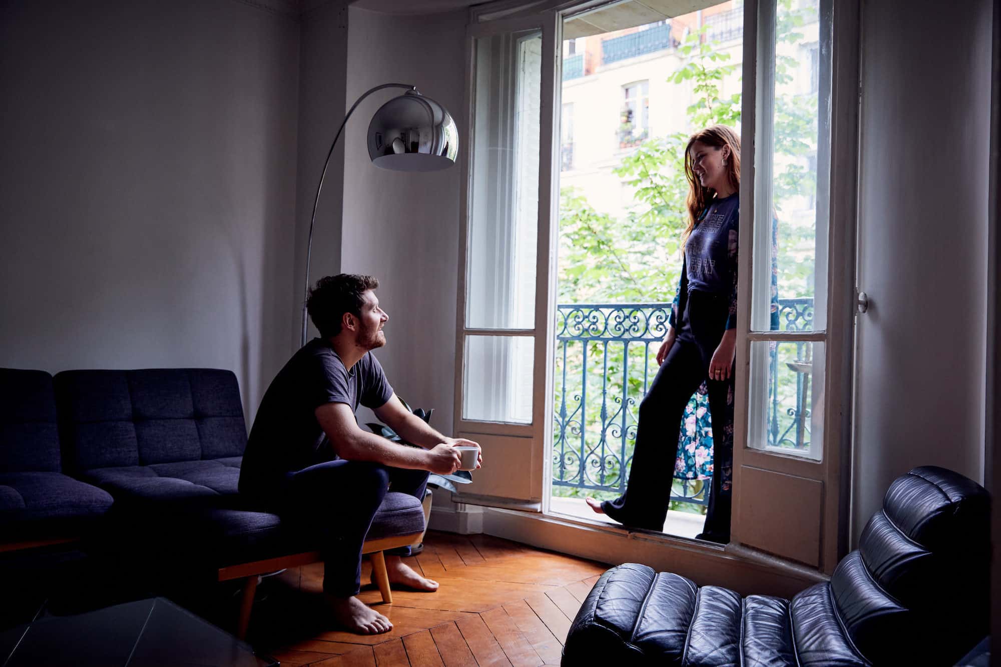 A young man and woman living in Paris and talking in their apartment which has wooden floors and a window that opens onto a balcony.