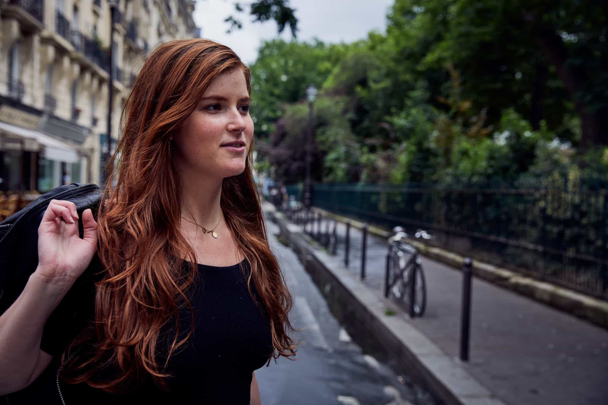 A young red-haired woman with a jacked slung over her right shoulder who loves living in Paris.