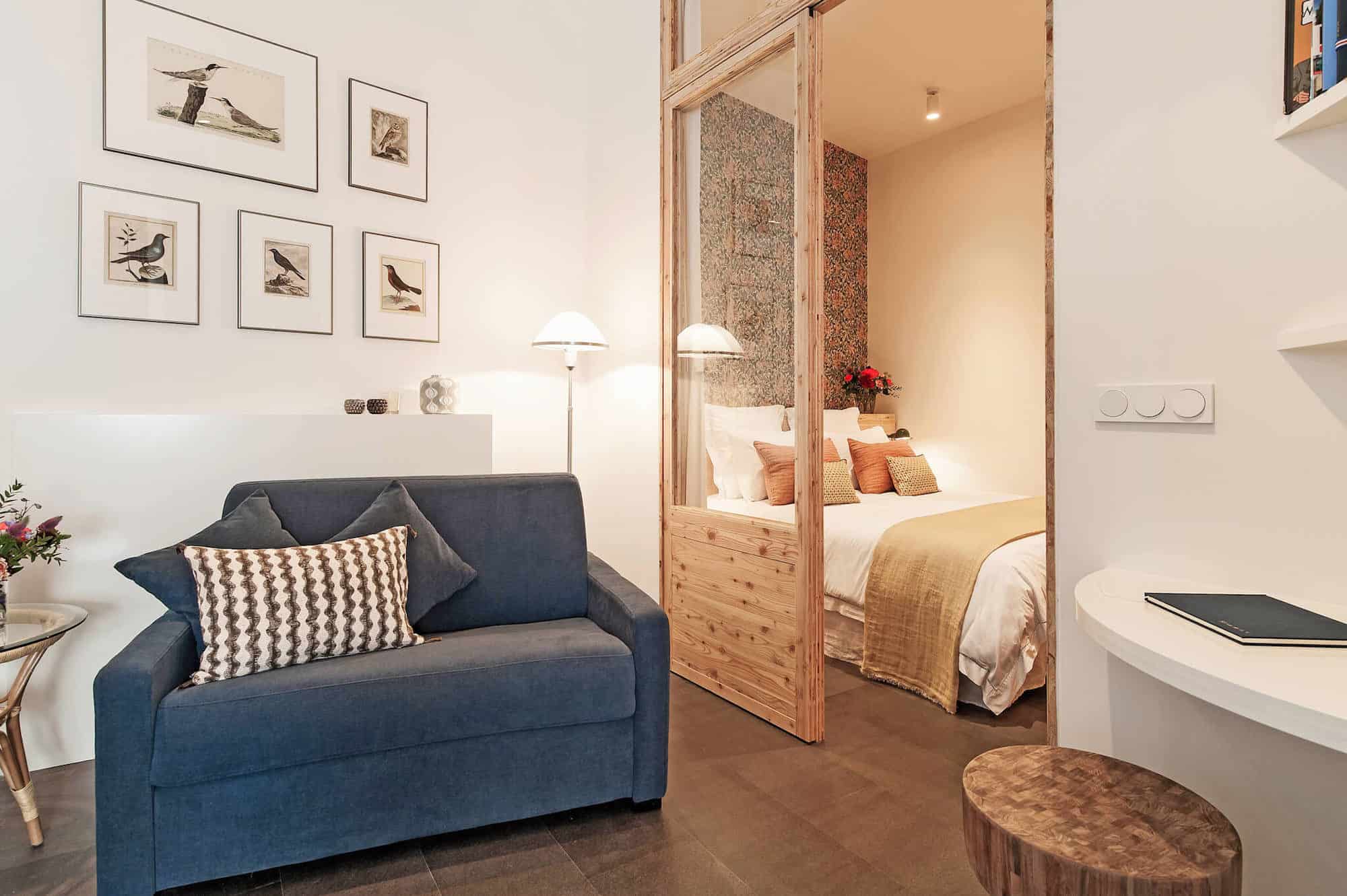 A beautiful rental apartment in Paris' Hotel de Ville area with a blue armchair, bird pictures up on the walls and a cozy double bed in the bedroom.