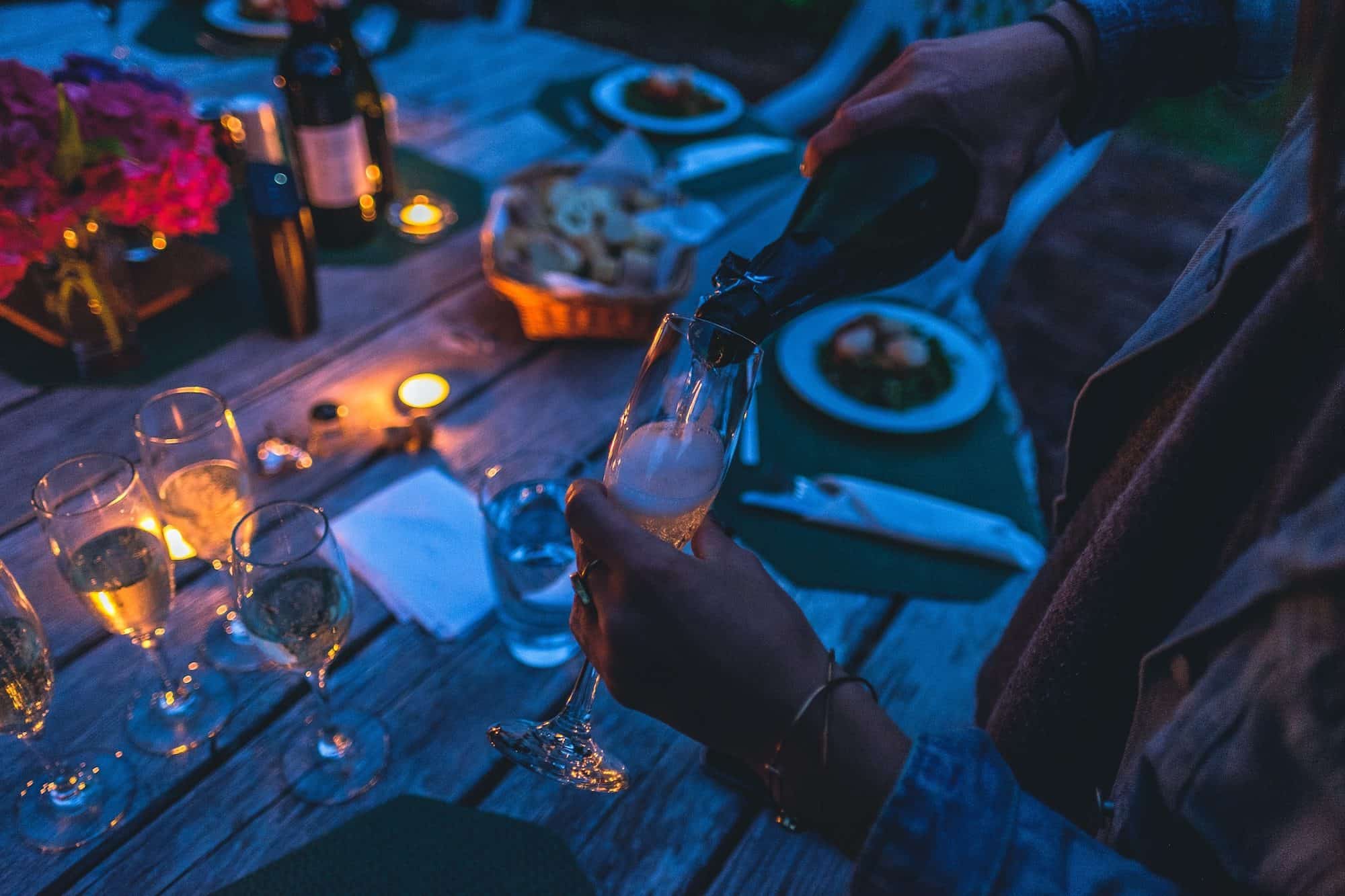 A woman pouring a glass of champagne at an outdoor candle-lit dinner on New Year's Eve.