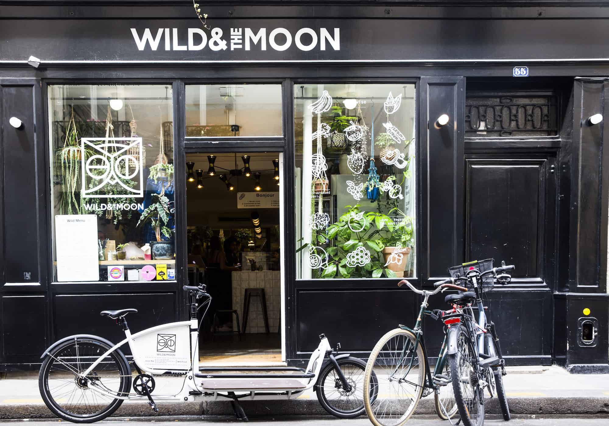 The window of Wild and the Moon gluten-free coffee shop in Paris with its black paneling and plants in the window and bikes parked outside.