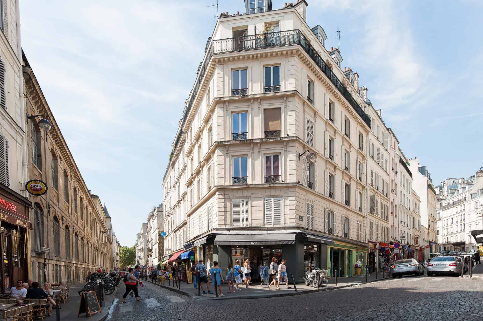 A street forking around  cream-colored iron-shaped building in Paris in summer, with people outside enjoying the sunshine. 
