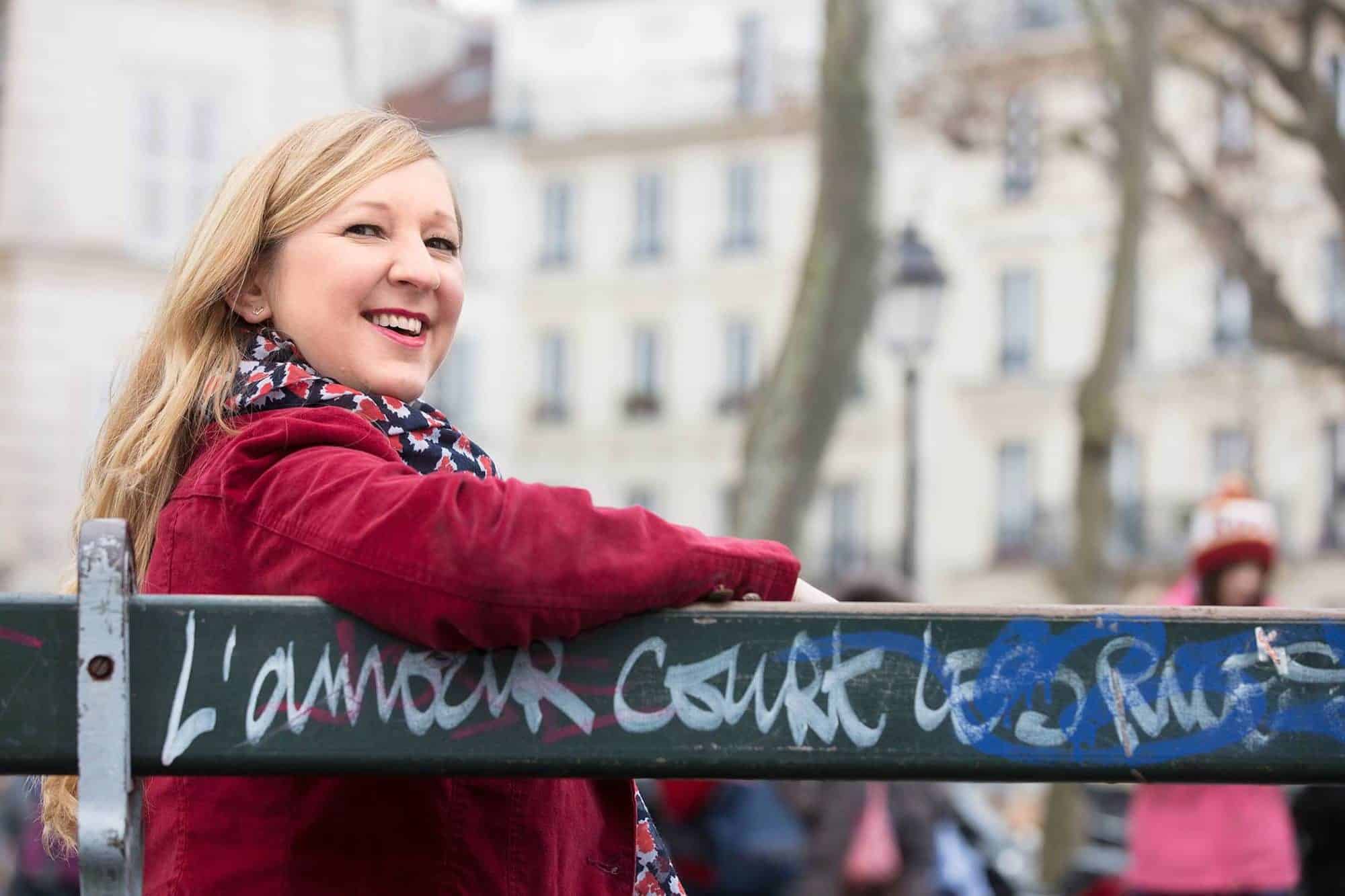 Expat blogger Lily Heise wearing a red jacket, sat on a Paris bench where the often-seen slogan 'Love runs through the streets' painted on it. She is smiling at the camera.
