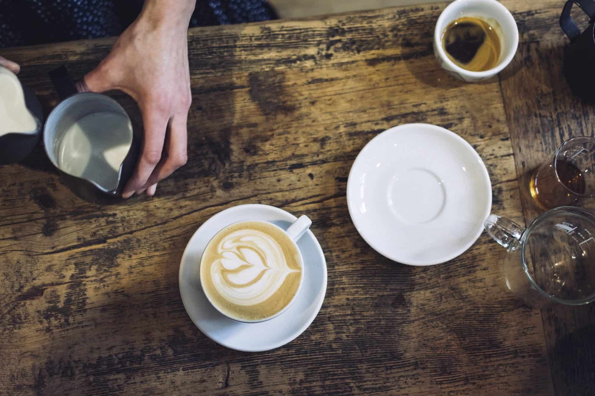 A barista making coffee in Paris, with a latte coffee with heart motif set down on a raw timber table.