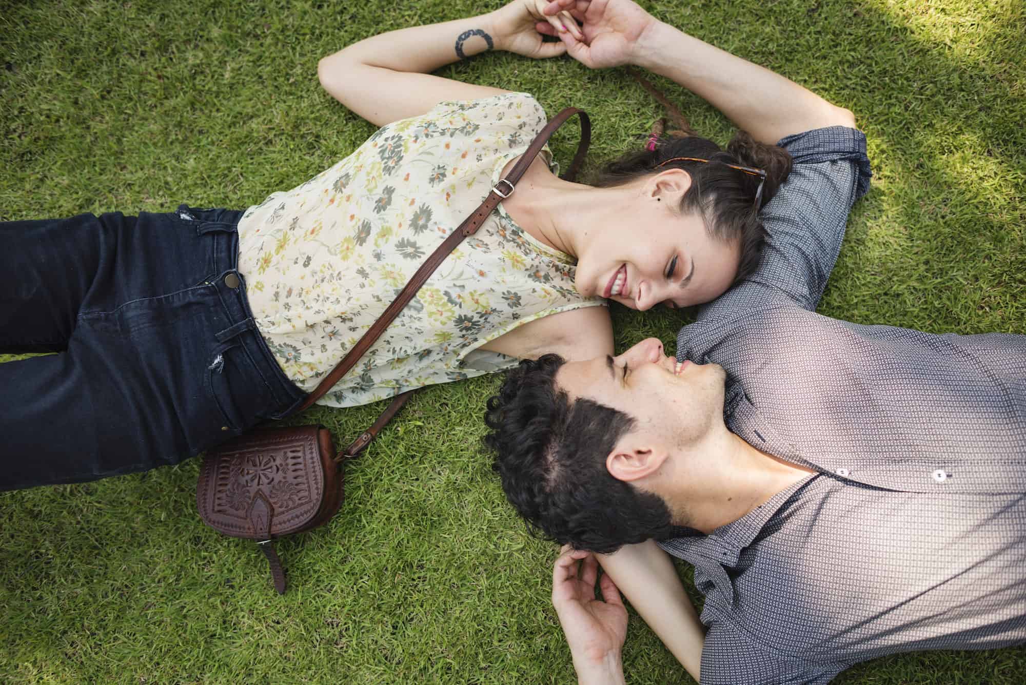 A couple holding hands and smiling at one another while lying on the grass.