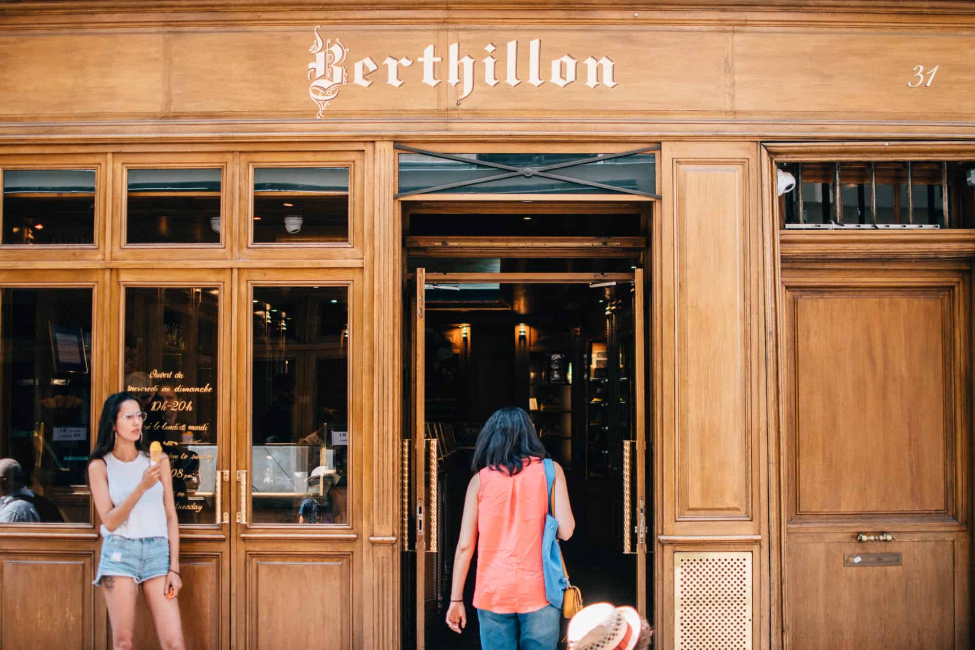The wood-paneled exterior of one Paris's Berthillon ice cream shops with a young woman standing outside, enjoying one of their soft ice creams.