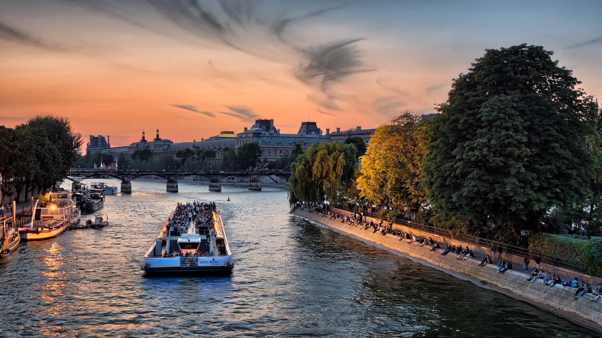 A view of the River Seine at sunset and a boat is sailing on the water, everything around it is golden from the sunlight.  