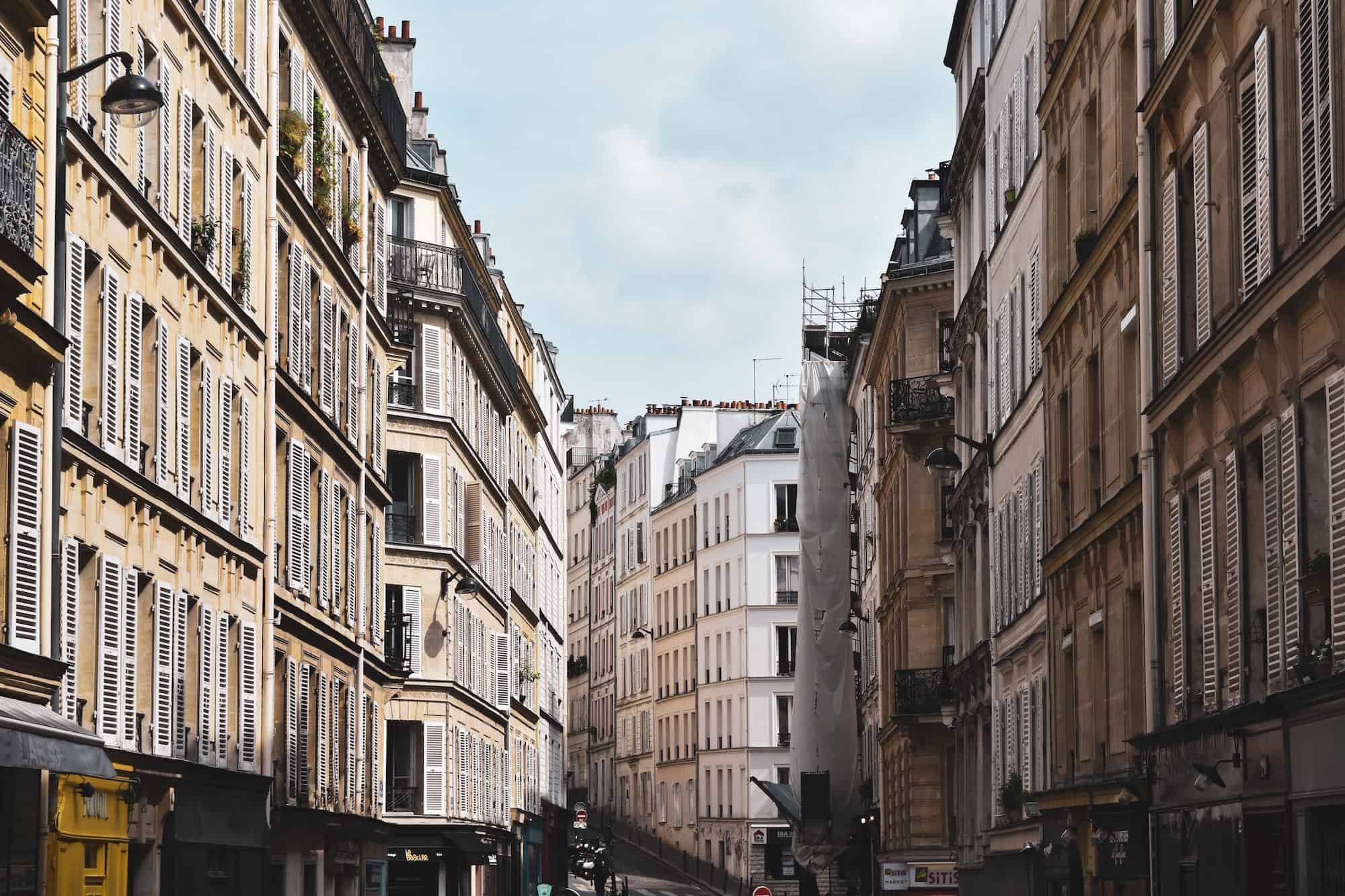 A view of white stone buildings with shutters lined up on both sides of a Paris street.