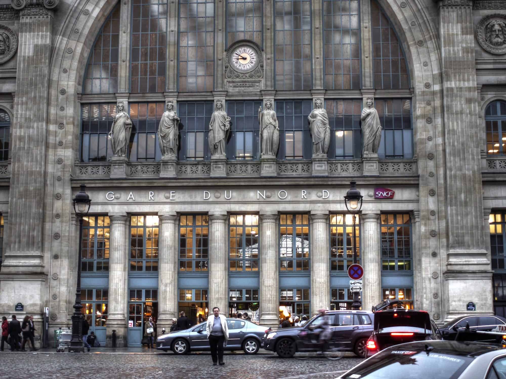 The beautiful statues lined up above the Gare du Nord's main entrance in Paris on a grey rainy day with a taxi driver standing in the foreground. 
