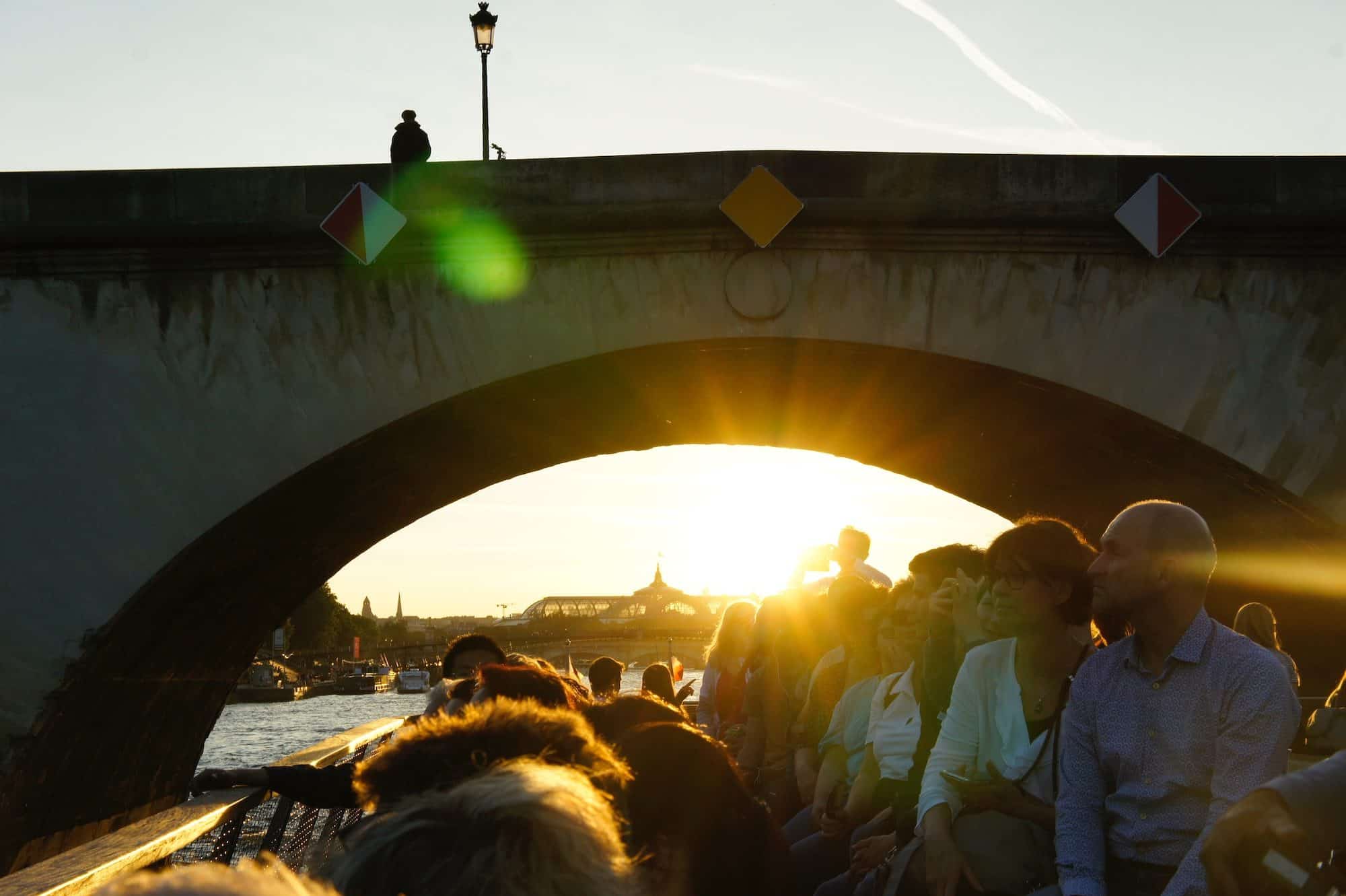 Tourists on a barge boat on the River Seine as the sun sets above the Grand Palais as seen peeping out from under one of the bridges straddling the river.