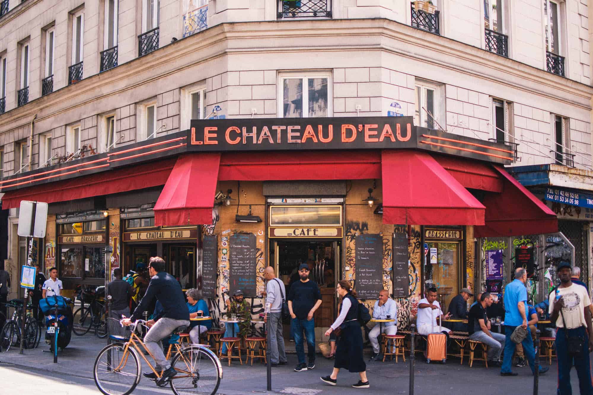 Outside a typical Parisian bistro called 'Le Chateau d'Eau' with a red awning and people sitting at tables outside.