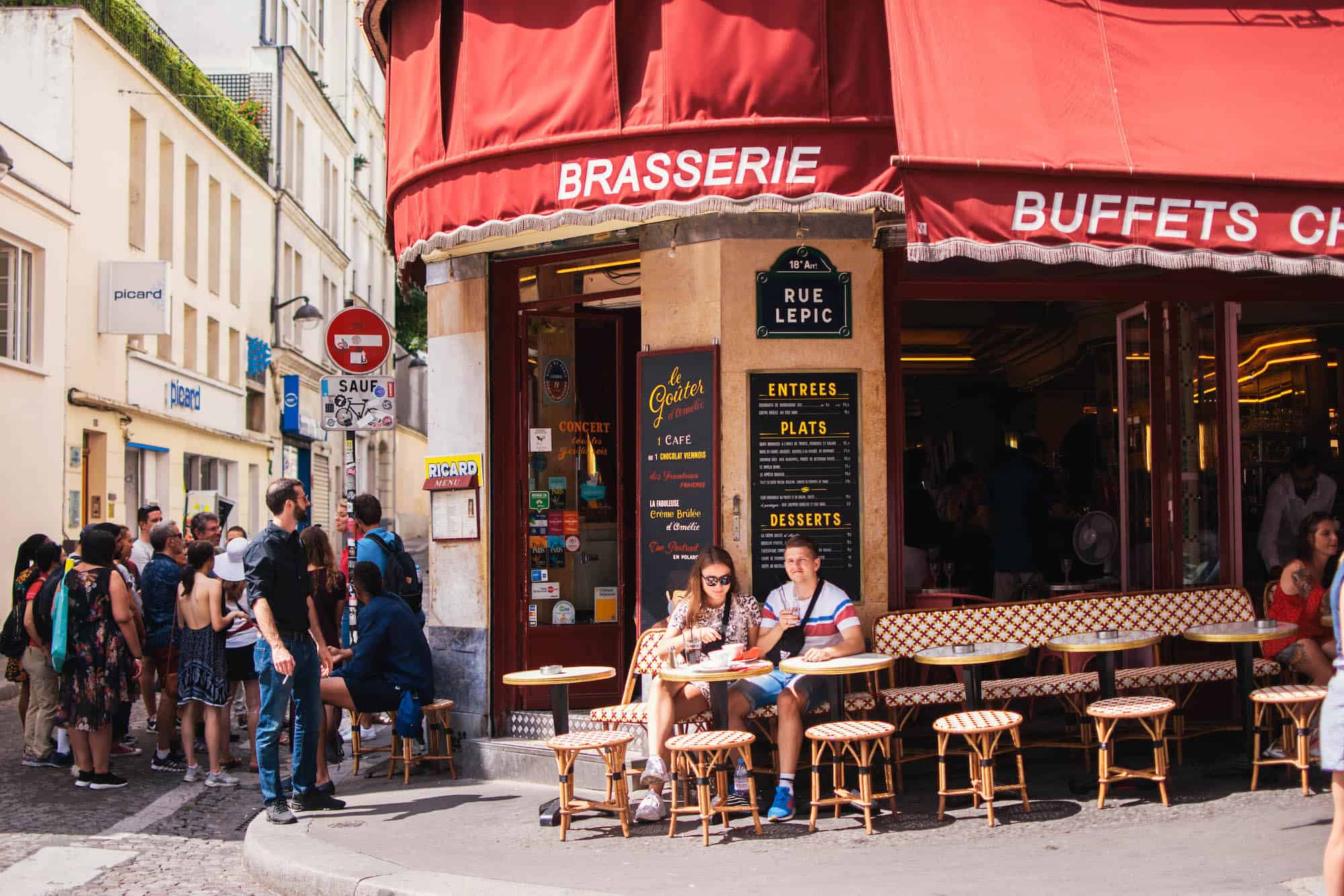 A typical Parisian brasserie with a red awning and people sitting on the terrace in the sunshine.