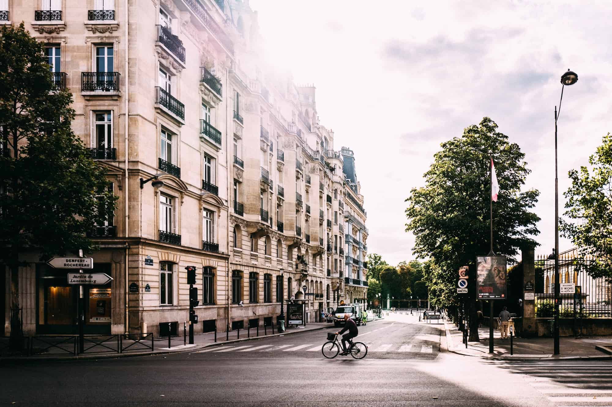 A cyclist rides across an empty street in Paris as the sun peeks from behind a classic Haussmann apartment building. 