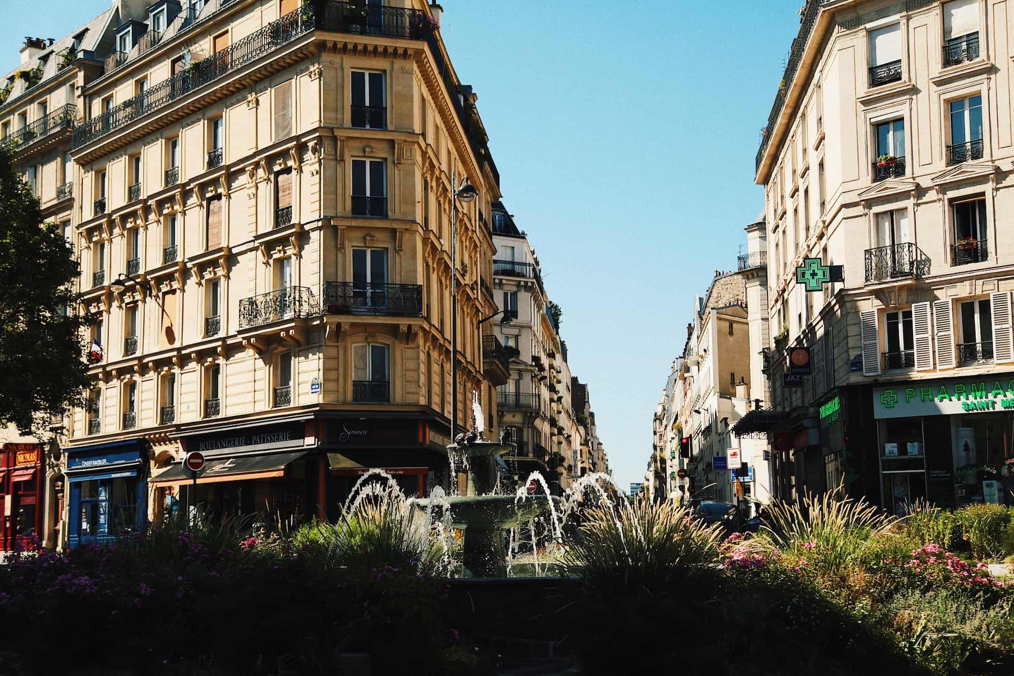 On a bright, sunny day, a fountain trickles in a Parisian square as the apartment buildings are bathed in sunlight. 