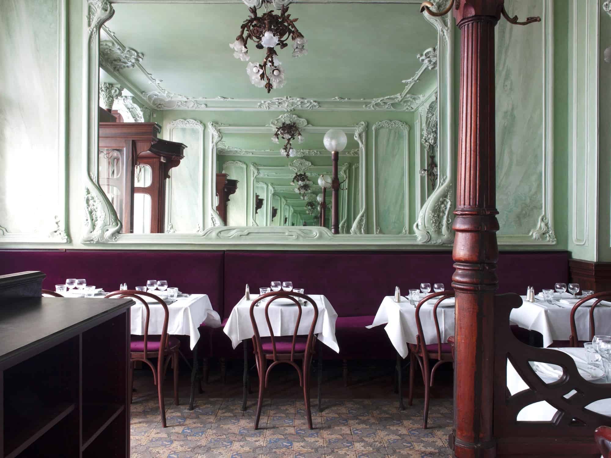 The art nouveau interior of Bouillon Julien restaurant in Paris. Pale green walls with a large mirror in the middle, chandeliers, maroon bench seat and chairs with tables with white table cloths and wine glasses.