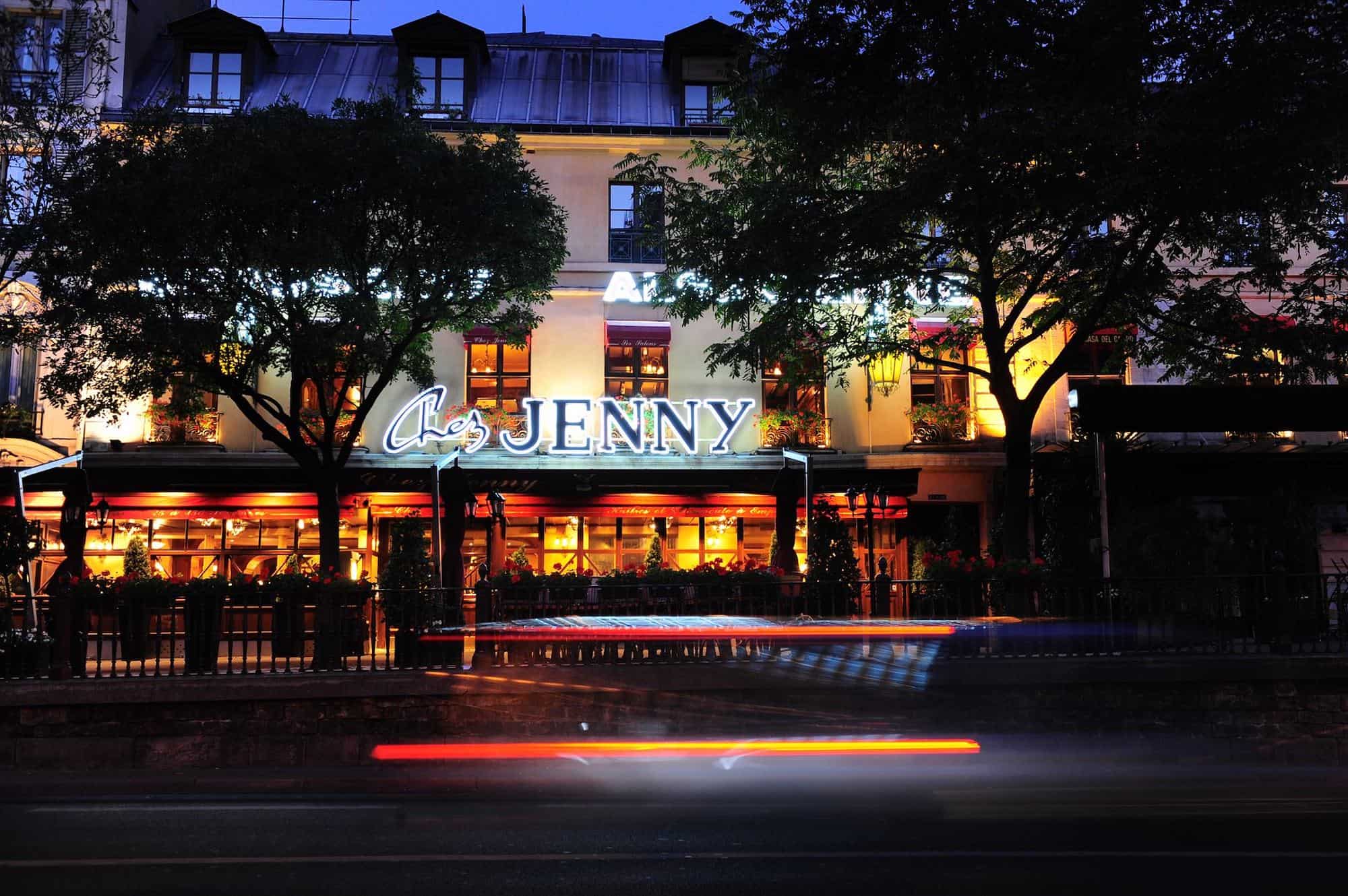 The exterior of the restaurant Chez Jenny in Paris at night time, with a large white neon sign saying Chez Jenny and streaks of light in front from a passing car. 