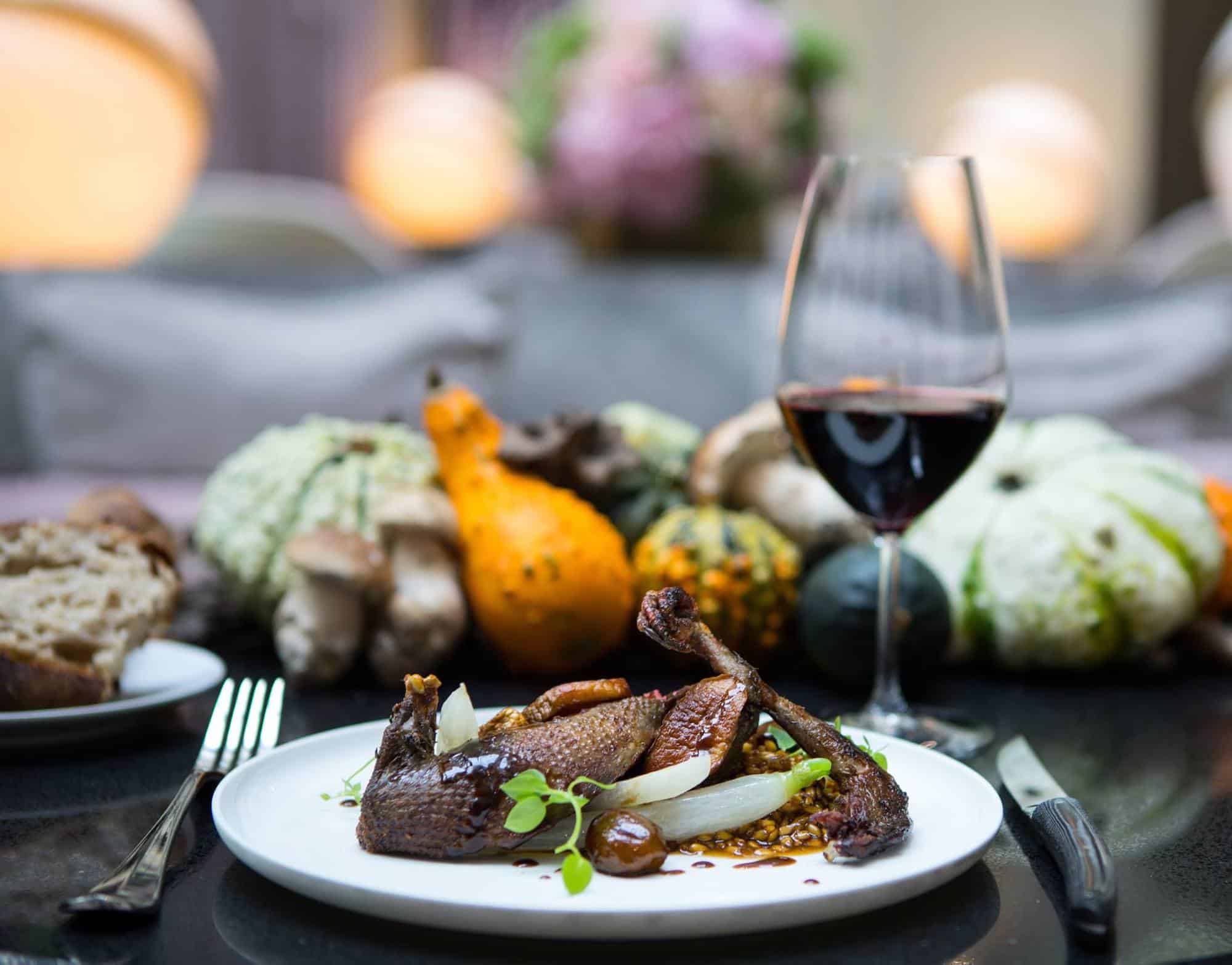 A white plate with roast pigeon, wheat risotto, turnip and chestnut, with cutlery on either side and a glass of red wine in the background, some bread, and an assortment of decorative squash.