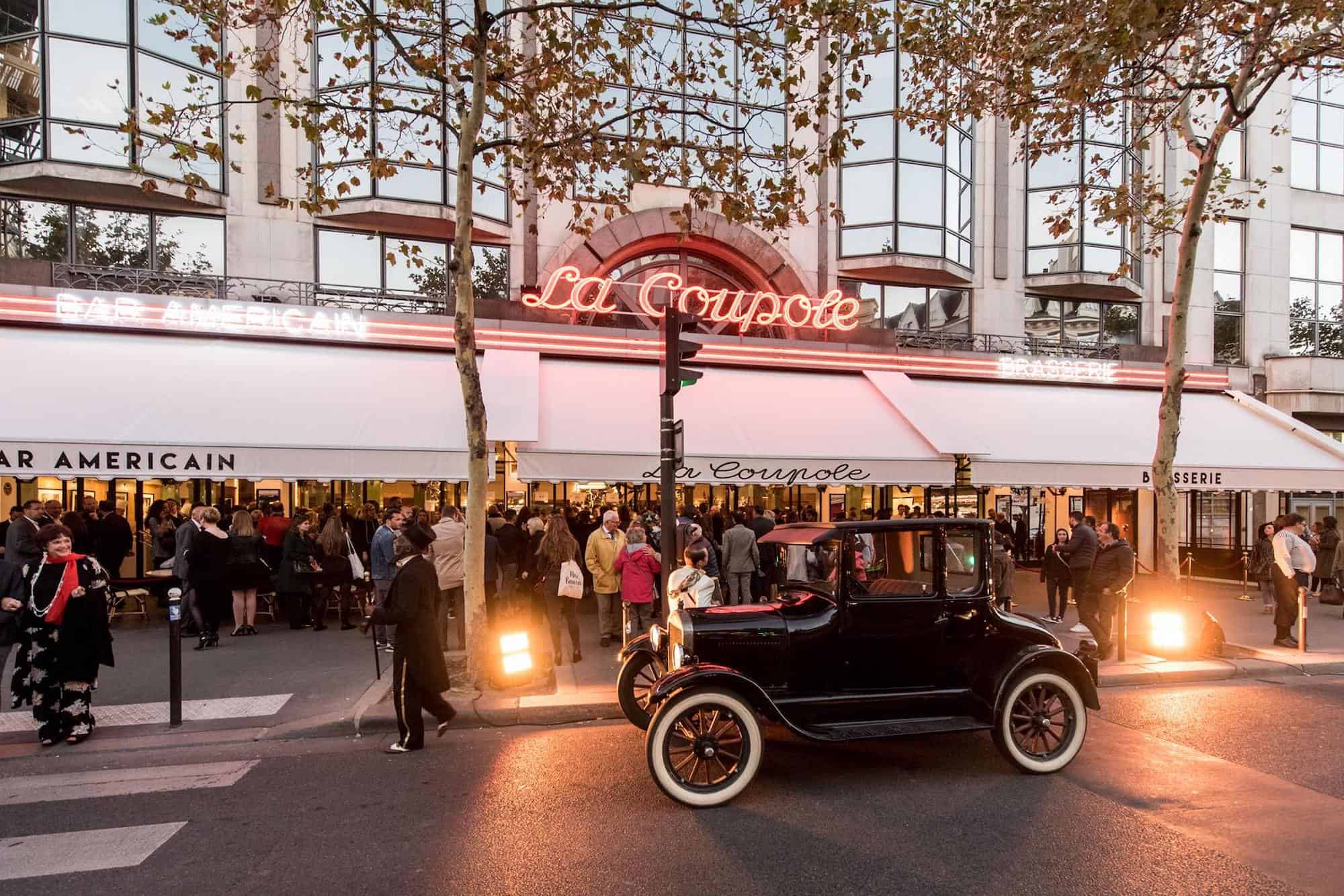 The exterior of La Coupole restaurant in Paris. A neon sign saying La Coupole, a white terrace shade, people standing in the terrace and an old fashioned black car parked out the front.