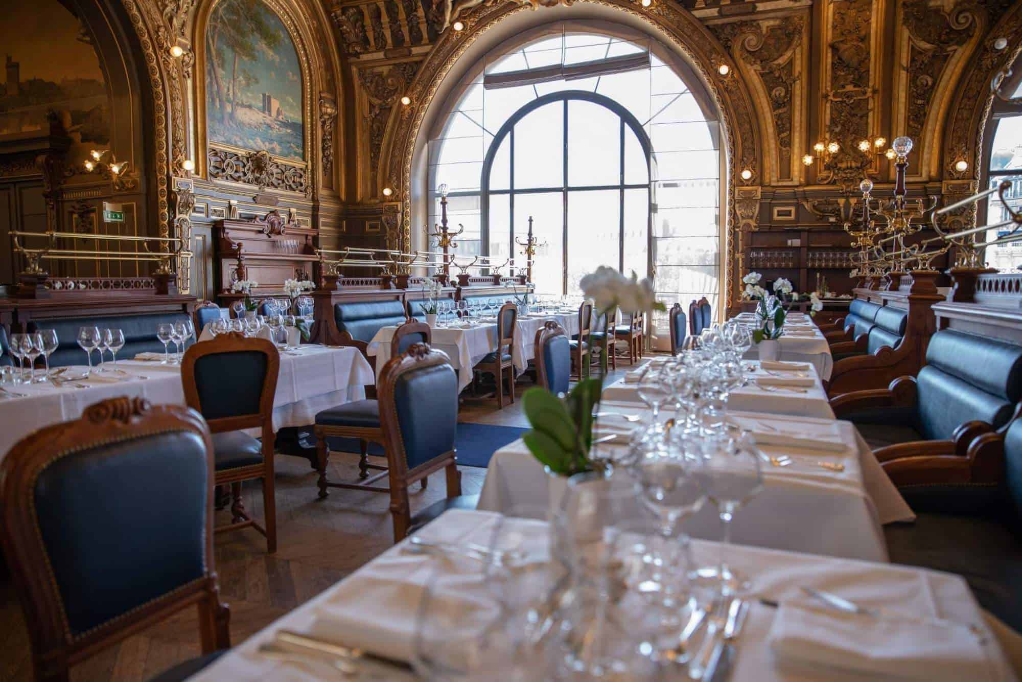 The opulent interior of the restaurant Le Train Bleu in Paris, tables covered in white tablecloths with white serviettes and wine glasses, blue upholstered seats, the walls have painted frescoes and carved gold detailing.