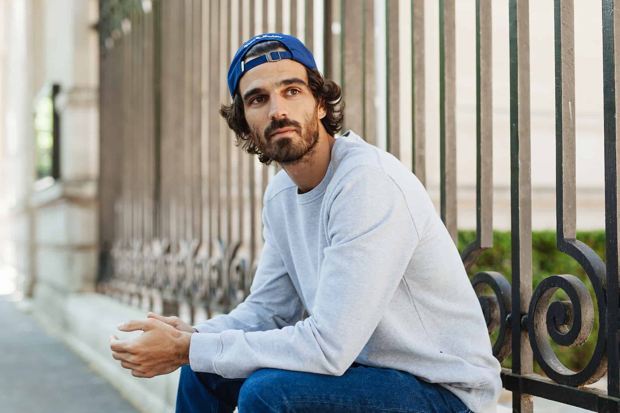 Sitting against an elegant wrought-iron gate, a model gazes off into the distance wearing a comfy grey sweatshirt by the ethical fashion brand forlife., and his wild brown curls are tamed by a deep blue baseball cap worn backwards.