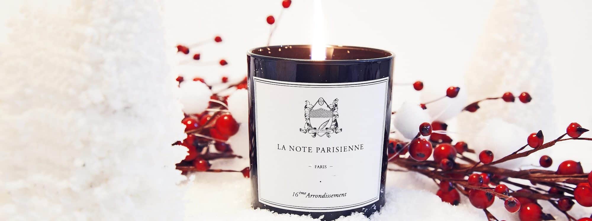 A lit candle from La Note Parisienne in a dark glass container with a black and white label. It is sat within a snowy, white Christmas setting with red berry sprays just behind it. 