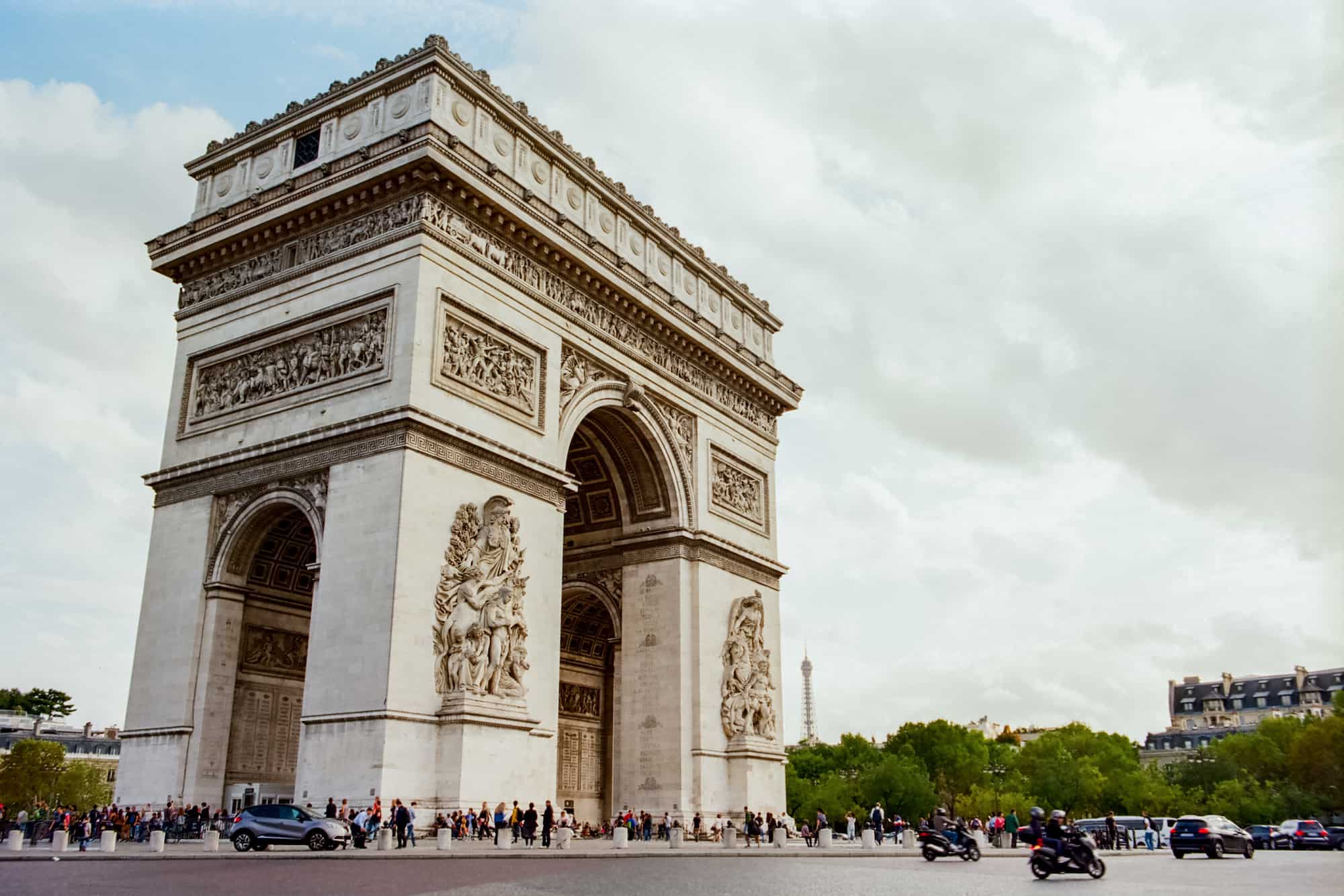 The Arc de Triomphe stands regally at sunset with the Eiffel Tower peeking in the background. Visitors mill about at the base, as motorists zip through the traffic circle. 