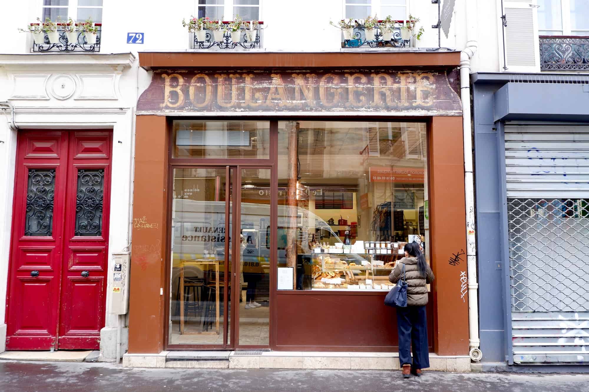 The facade of a boulangerie. There is a vintage sign above the windows. A woman is looking in the windows at the pastries on display.