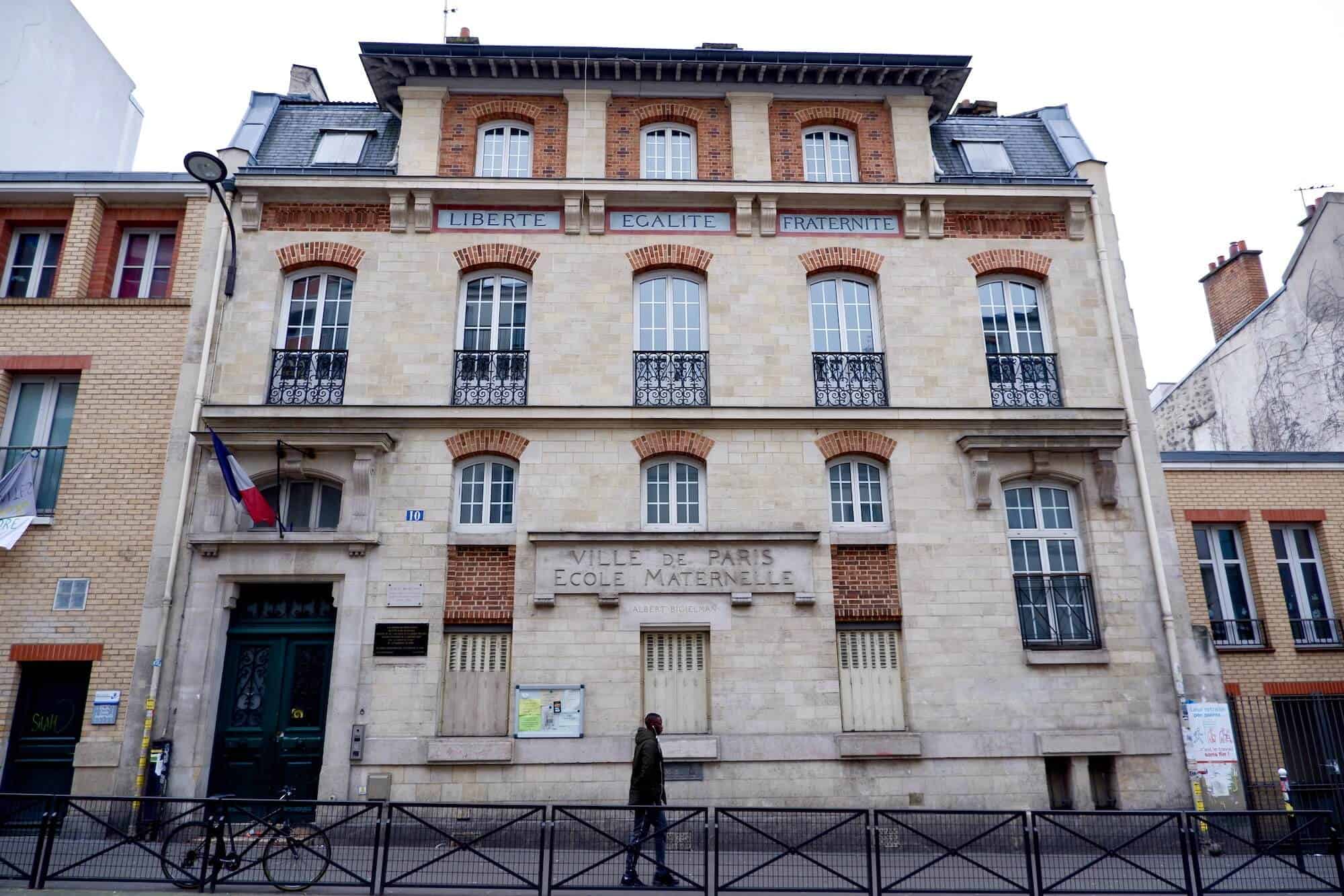 The facade of a kindergarten. The words 'Liberte, Egalite, Fraternite' are at the top of the building, lower down are the words 'Ville de Paris, Ecole Maternelle'. There is a French flag flying above the front door. There is a man walking past.