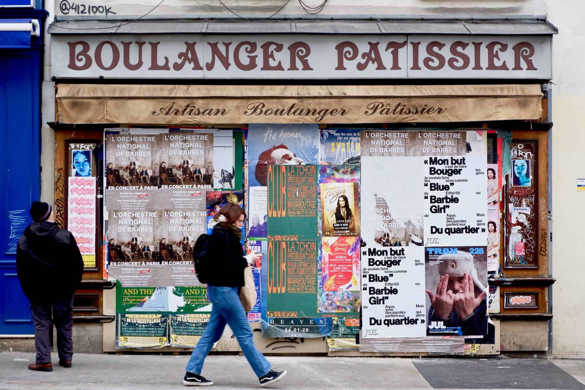 An old boulanger/patissier that has been covered in promotional music posters. An elderly man to the left is looking up at the posters while a young woman is walking past, slightly blurred.