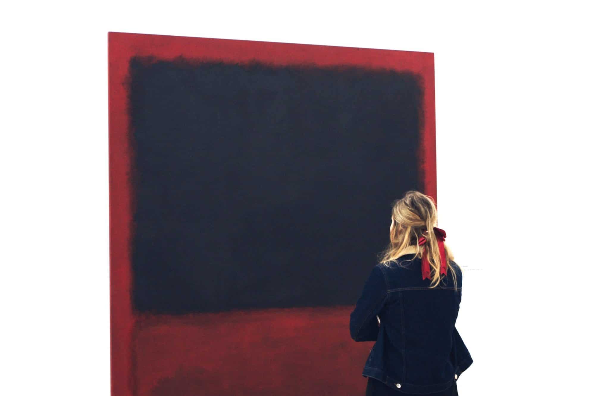 A blonde woman standing infant of a Rothko painting. She's wearing a denim jacket and has a red ribbon in her hair. The Rothko painting is dark red with a black square.