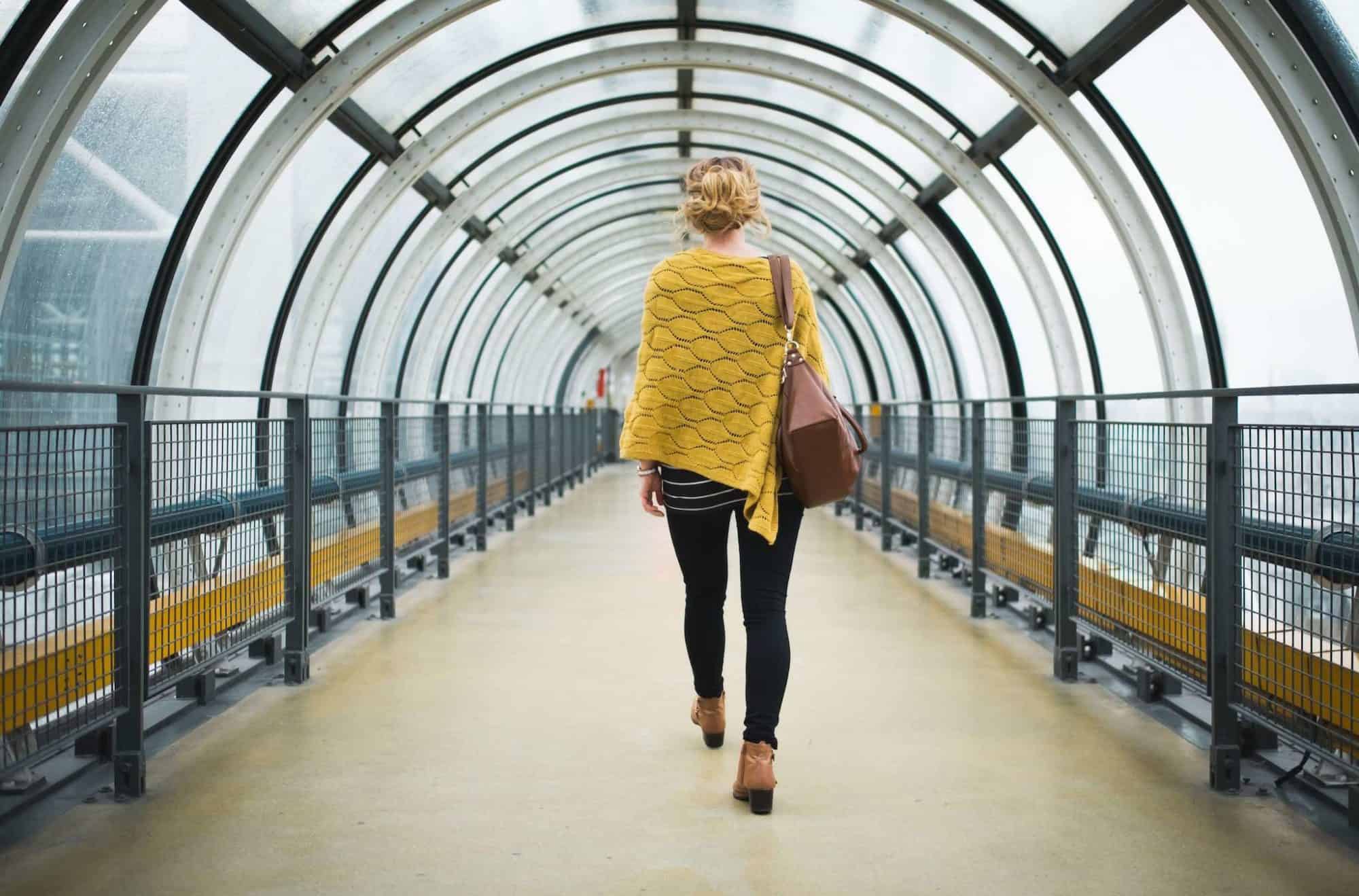 A blonde woman wearing a yellow poncho, black jeans, and brown boots and handbag is walking down a glass cylindrical corridor in Centre Pompidou. The photo is taken from behind. 