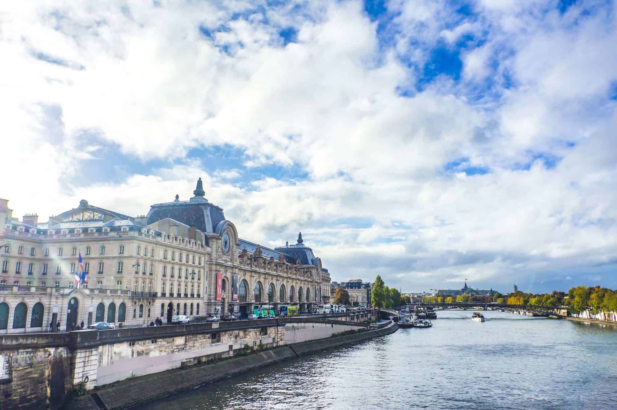 The outside of the D'Orsay Museum, on the Seine river in Paris. The sky is blue with some clouds, the sun is shining, the trees along the river are green. There is a boat going down the river.