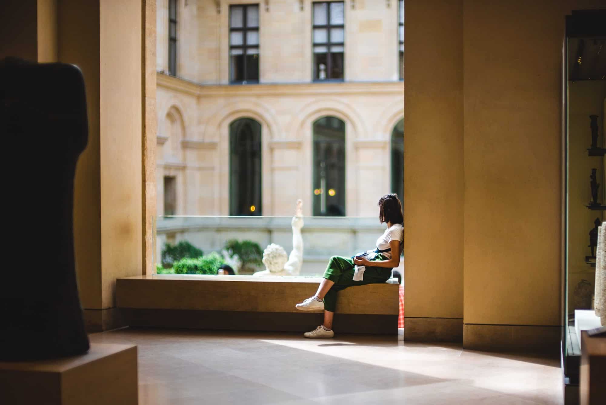 A brunette girl in a white t-shirt and green pants, wearing white sneakers, sitting on a bench seat in the Louvre Museum. She is looking away from the camera to an inner sculpture courtyard.