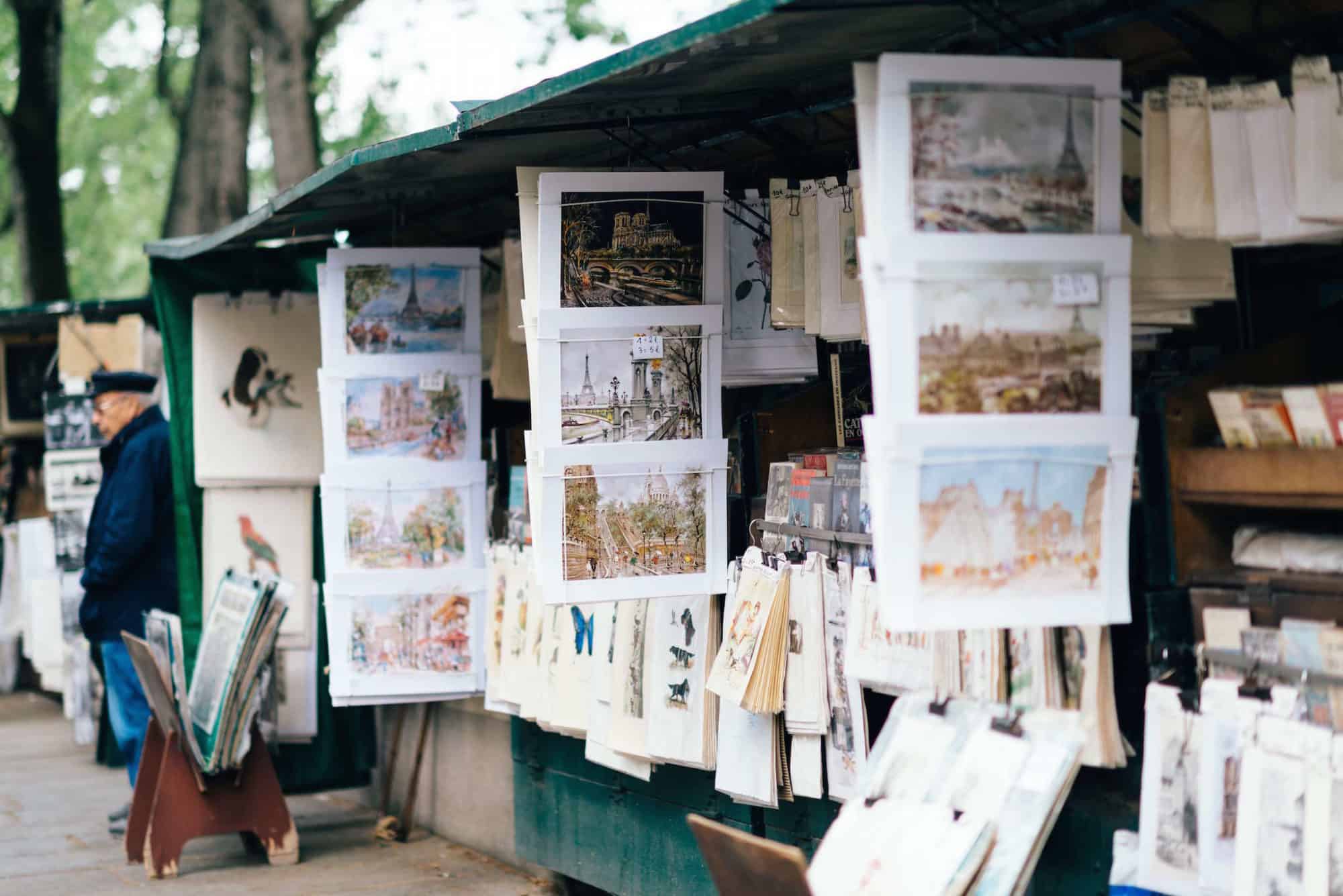 An old man stands by stalls of art near the Seine in Pairs. Beautiful paintings that display scenes of Paris can be seen hanging from the stalls.