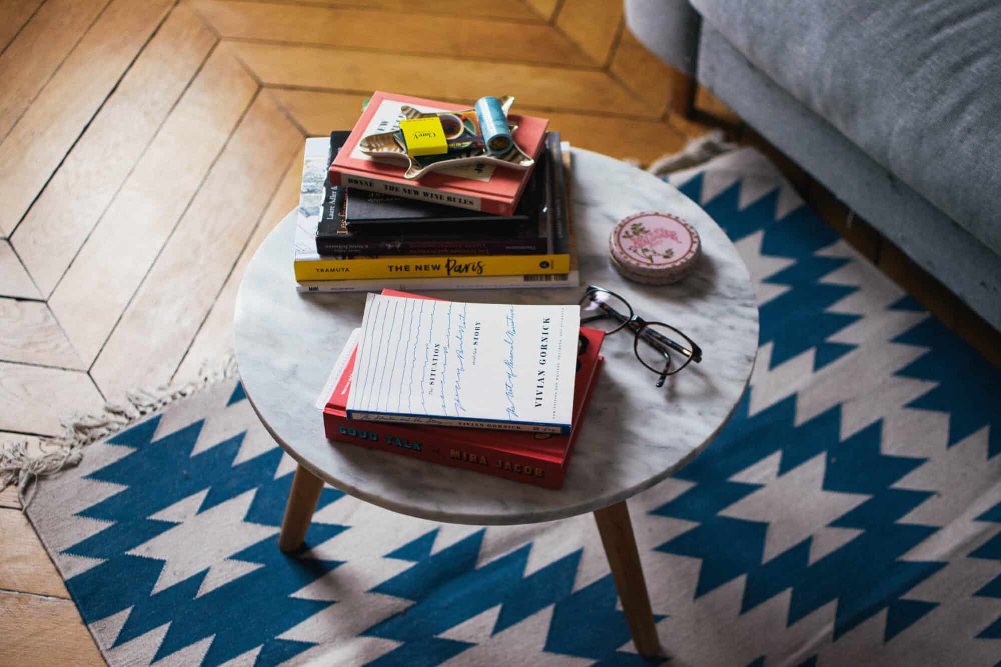 Uma mesa de mármore ao lado de um sofá cinza em um apartamento em Paris, em cima há uma variedade de itens, incluindo livros e um par de óculos.