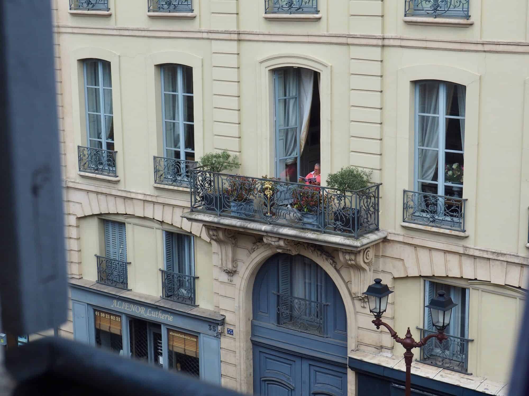A woman in a pink shirts reads on her balcony in Paris, with her large door open and plants next to her on the balcony.