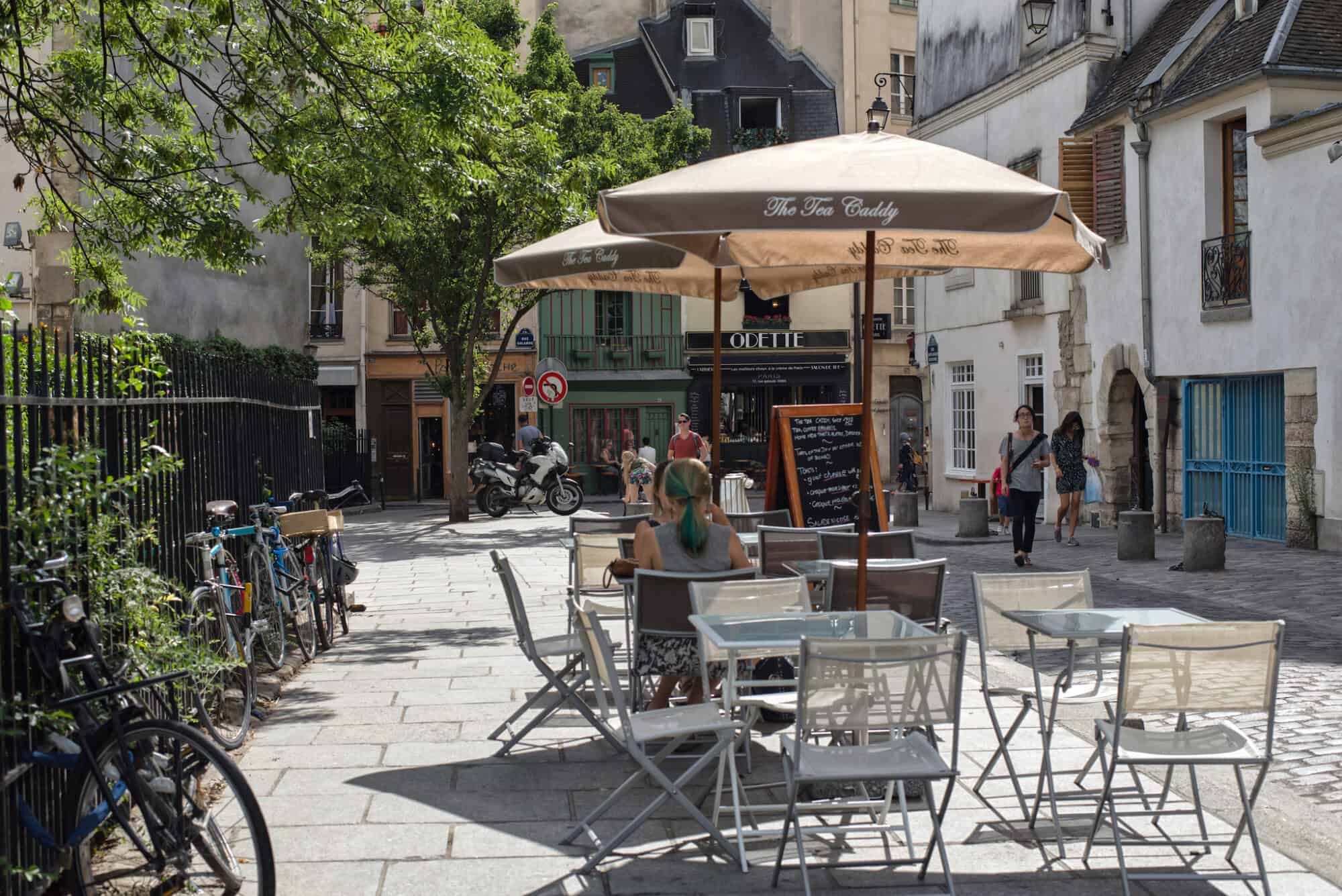 People sit at tables and chairs and stroll down a street in the left bank of Paris on a sunny day.
