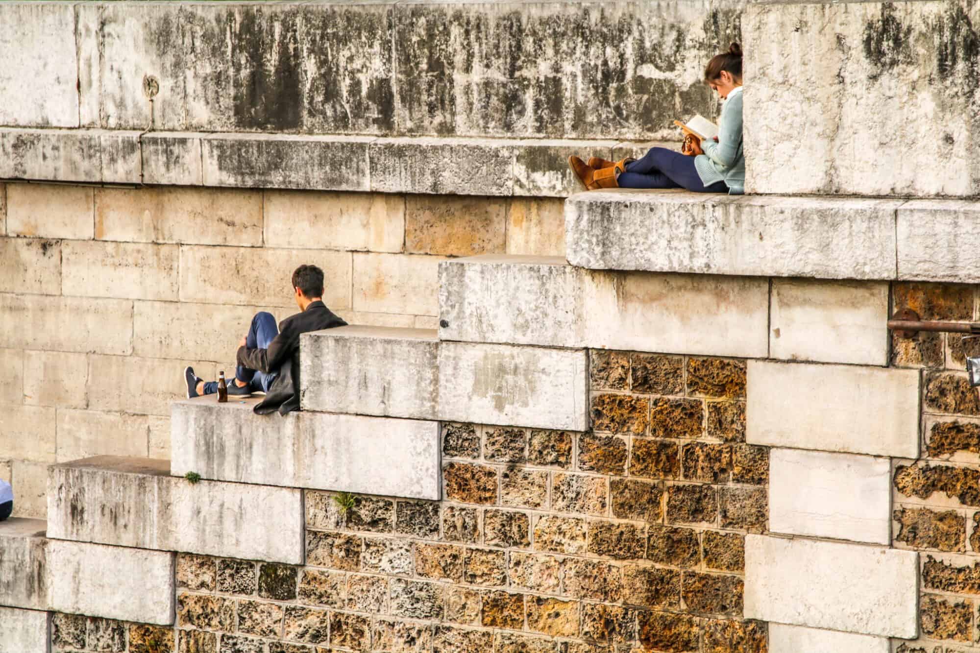 People are seen sitting on large stone steps near the Seine River in Paris; A man sits holding his leg and a woman a few steps above sits while reading a book.