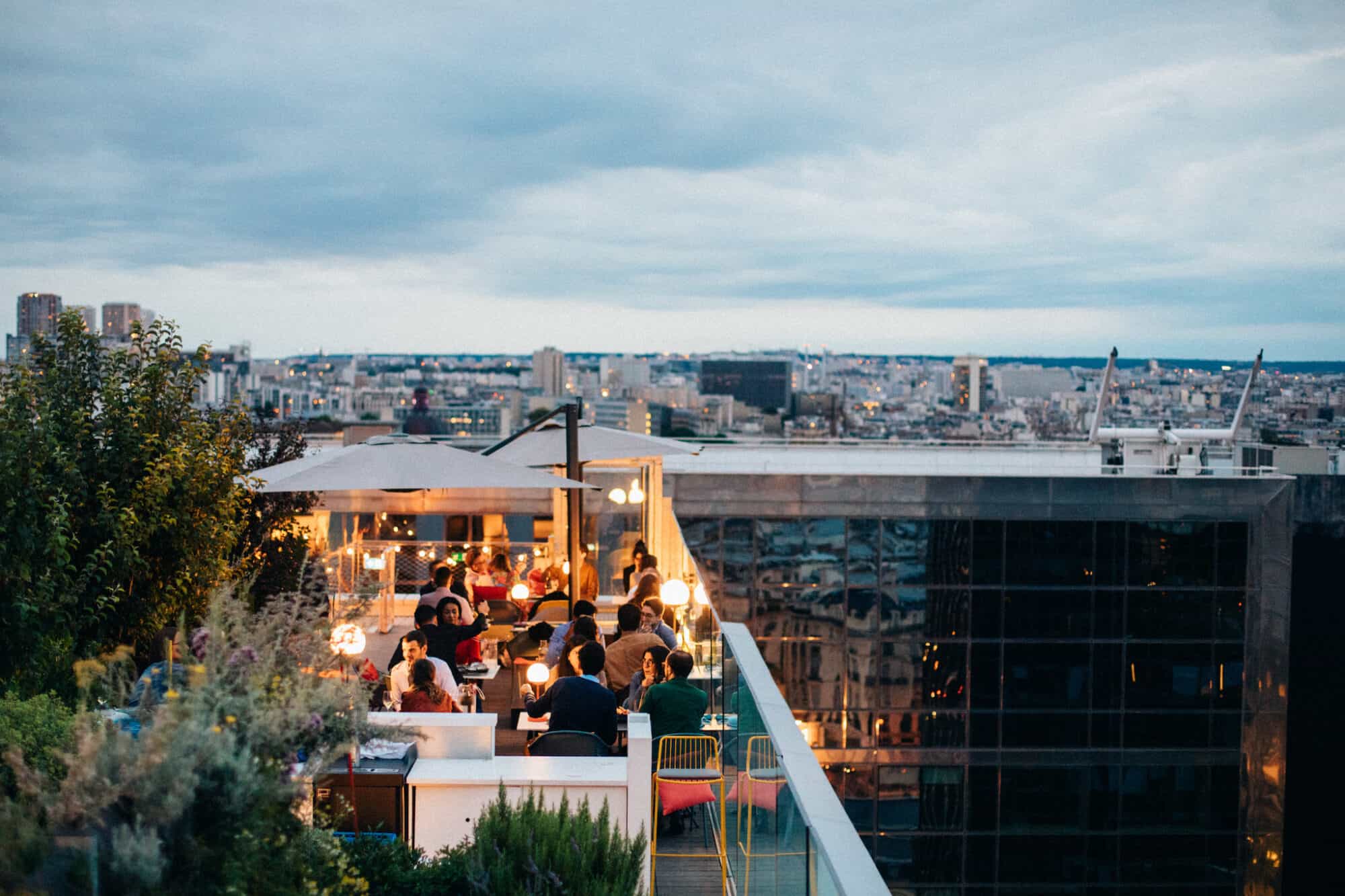 The terrace of the rooftop bar Laho. It is dusk and there are people seated at tables. The lights are on and they are seated underneath umbrellas. There are some trees and plants on the left. In the background you can see a view of Paris under a grey, cloudy sky.