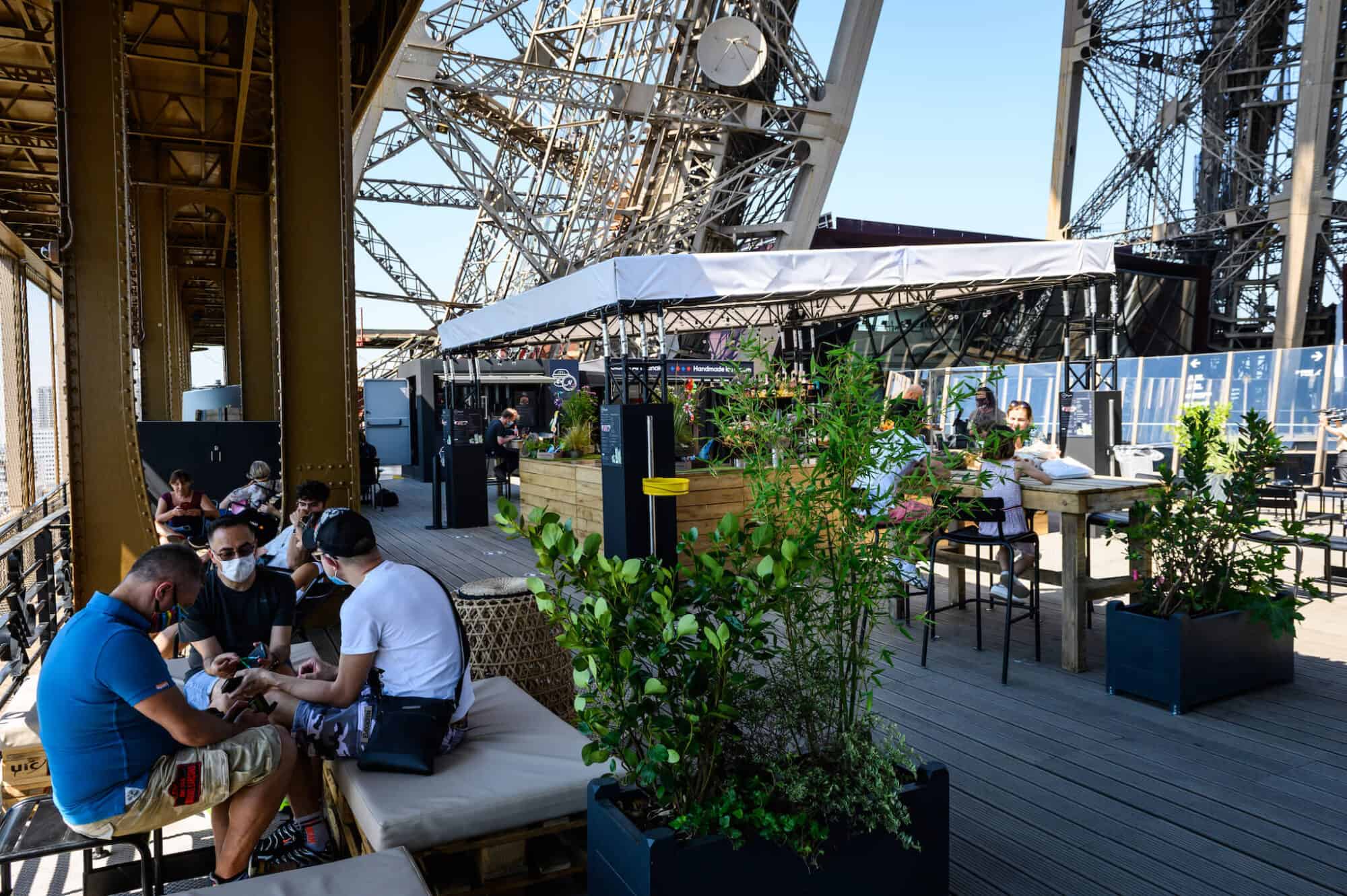 The wooden terrace of the Eiffel Tower in Paris. There are groups of people seated, some wearing face masks. You can see the pillars of the Eiffel Tower around them. There is a wooden bar in the middle with a white tarp roof. There are also some pot plants.