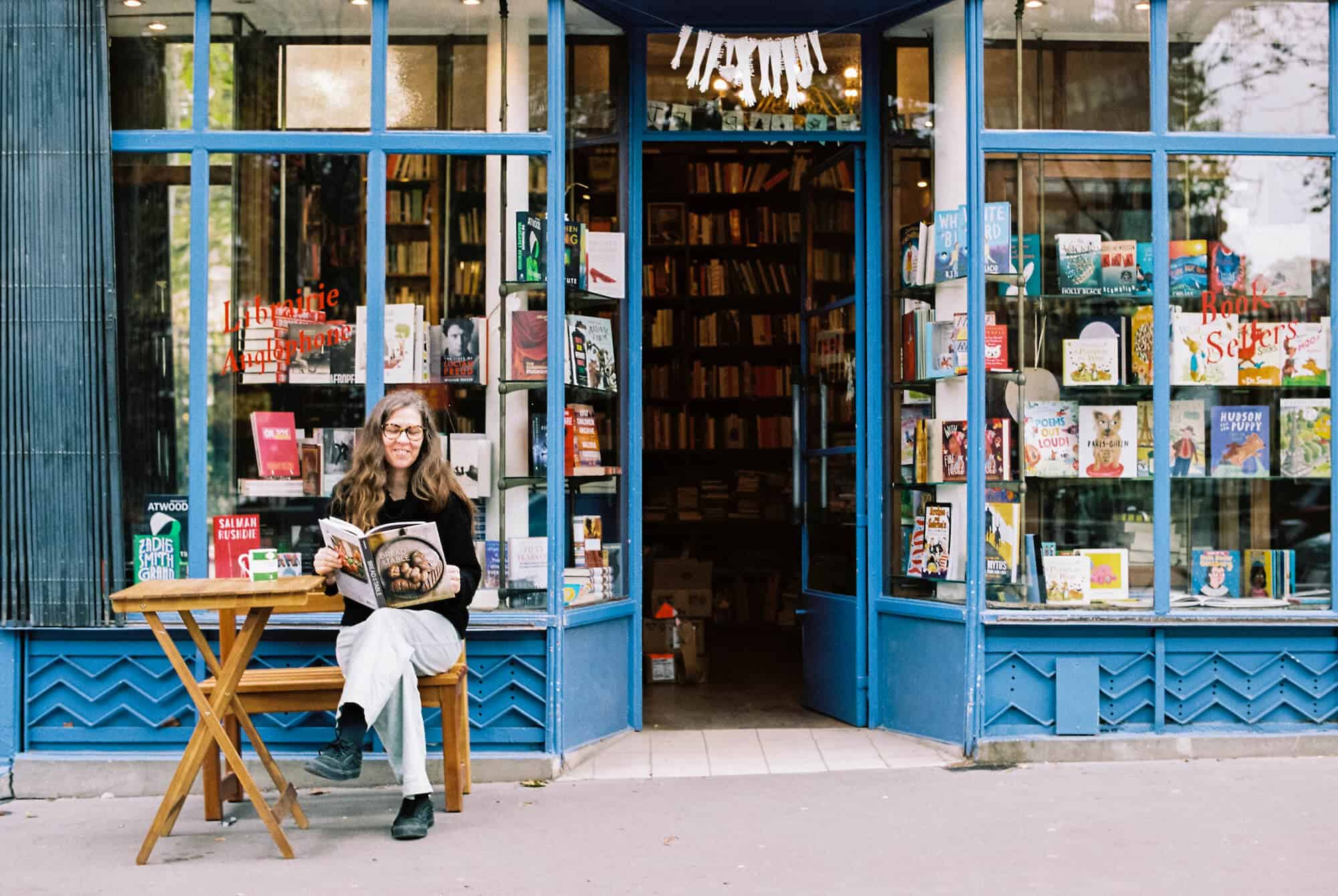 Penelope Fletcher, bookseller at the Red Wheelbarrow, sits outside the bright, blue and beloved bookstore with a magazine in her hands and smile on her face. 