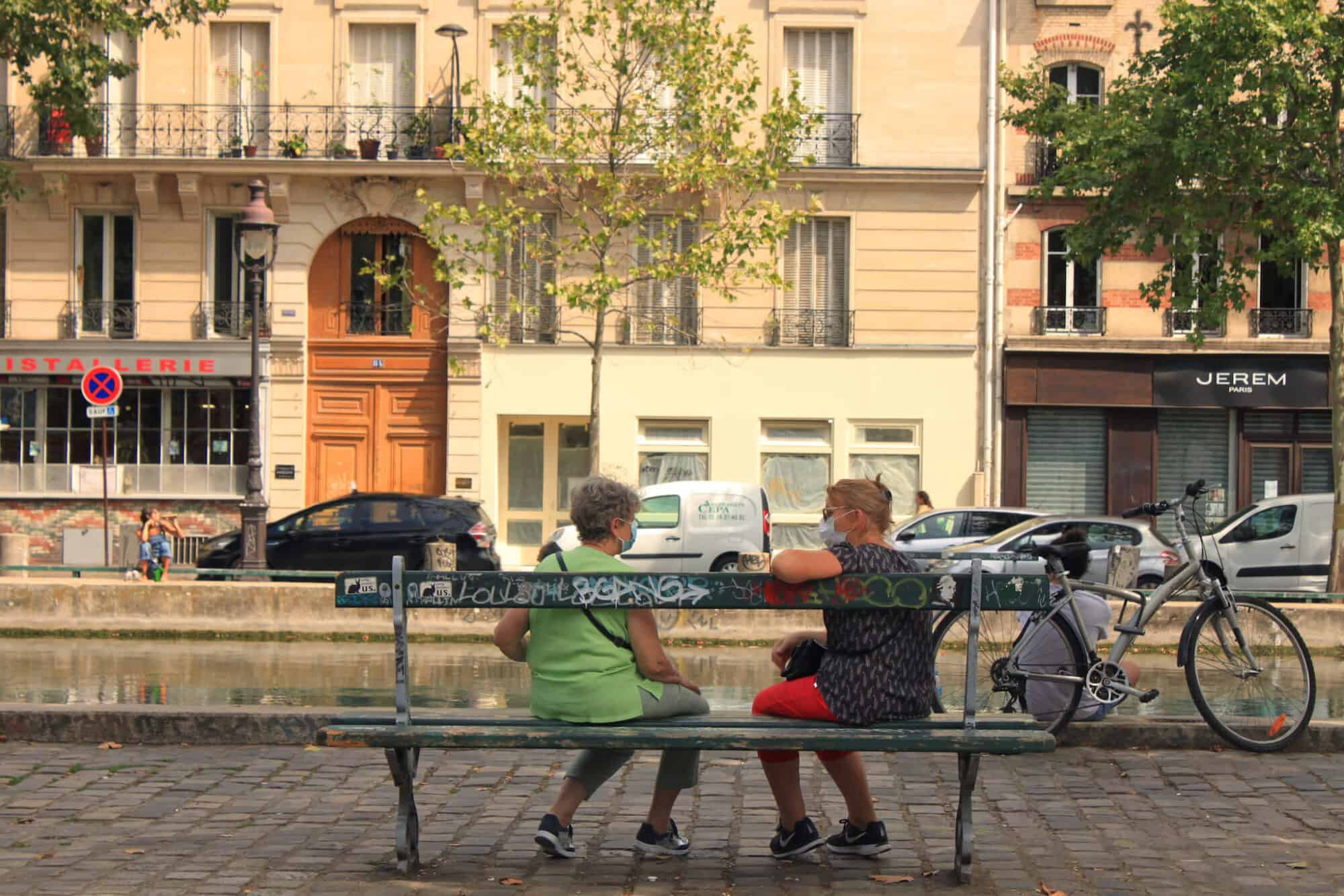 Two women sit on a bench by Paris's Canal St. Martin and speak to each other while wearing face masks.