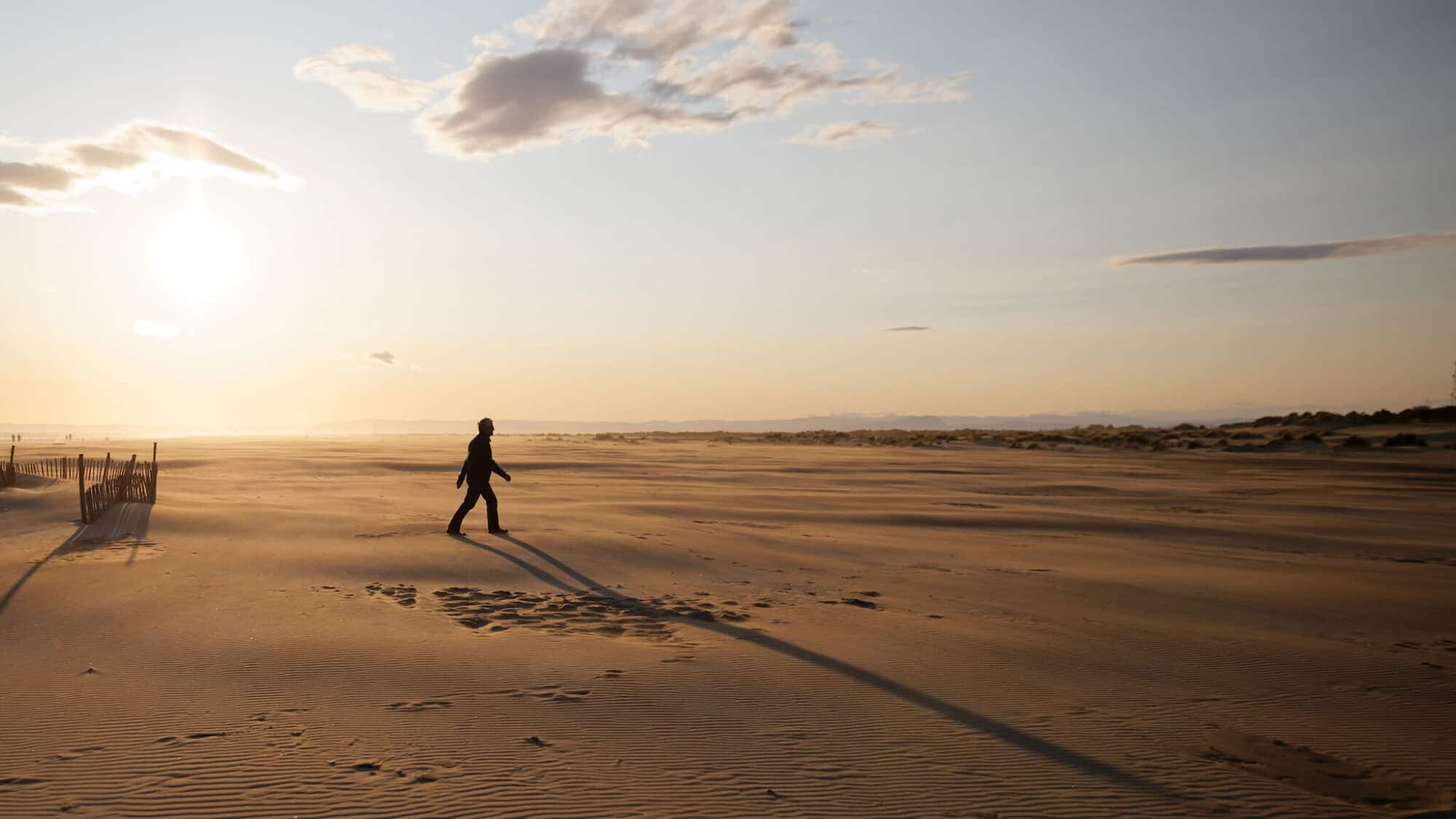 A person walks on the sandy beach of the Camargue at sunset, casting a shadow on the ground.